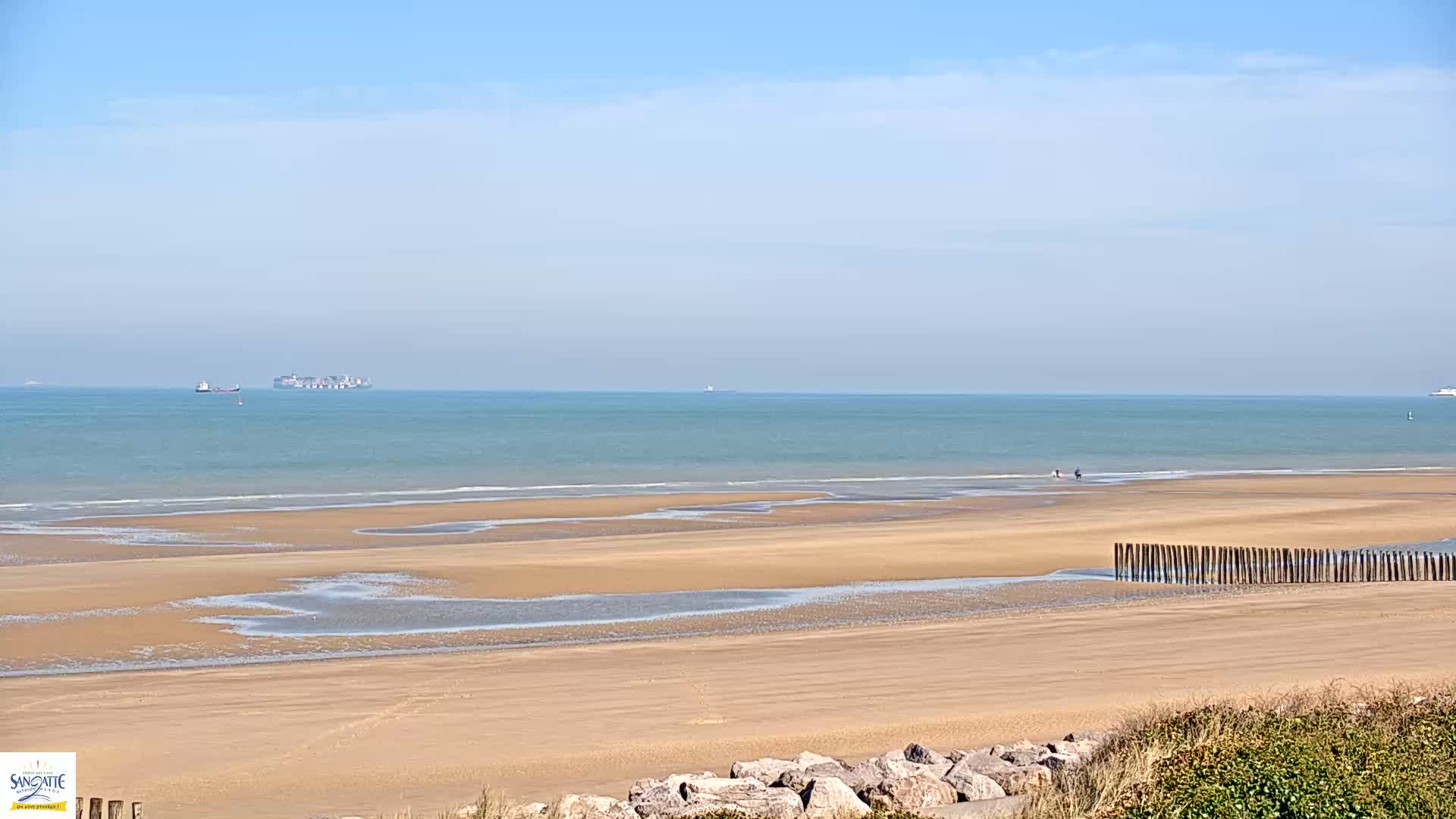 A wide sandy beach with calm, light-blue water and a few distant ships under a clear, bright sky stretches to the horizon.