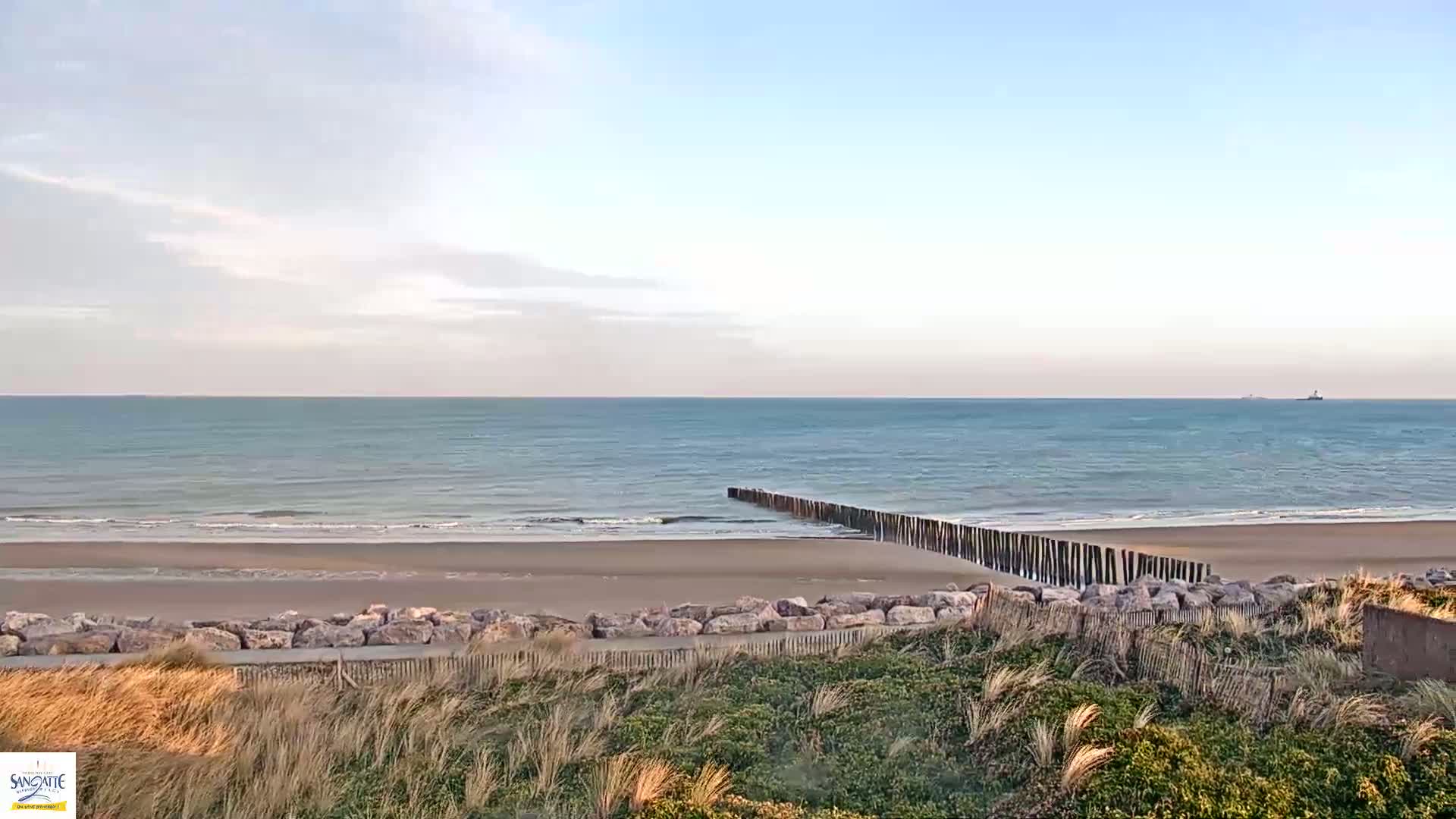 A sandy beach with calm ocean waves and a wooden breakwater is seen from a dune under a partly cloudy sky.