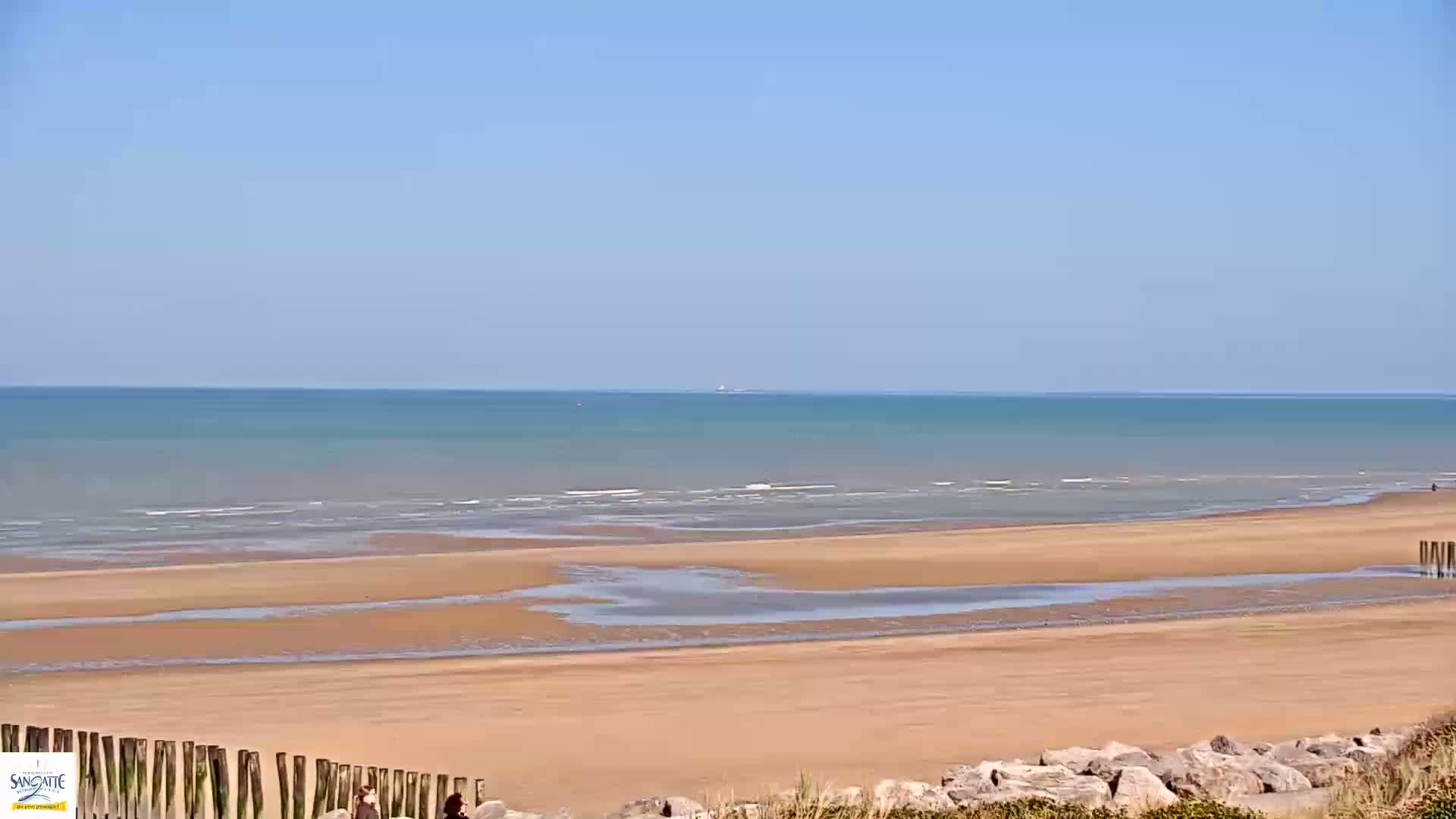 A wide sandy beach stretches to a calm, light-blue ocean under a clear blue sky.