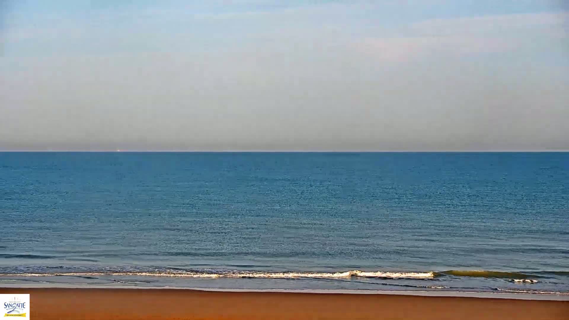 A calm, blue ocean meets a sandy beach under a pale, clear sky.