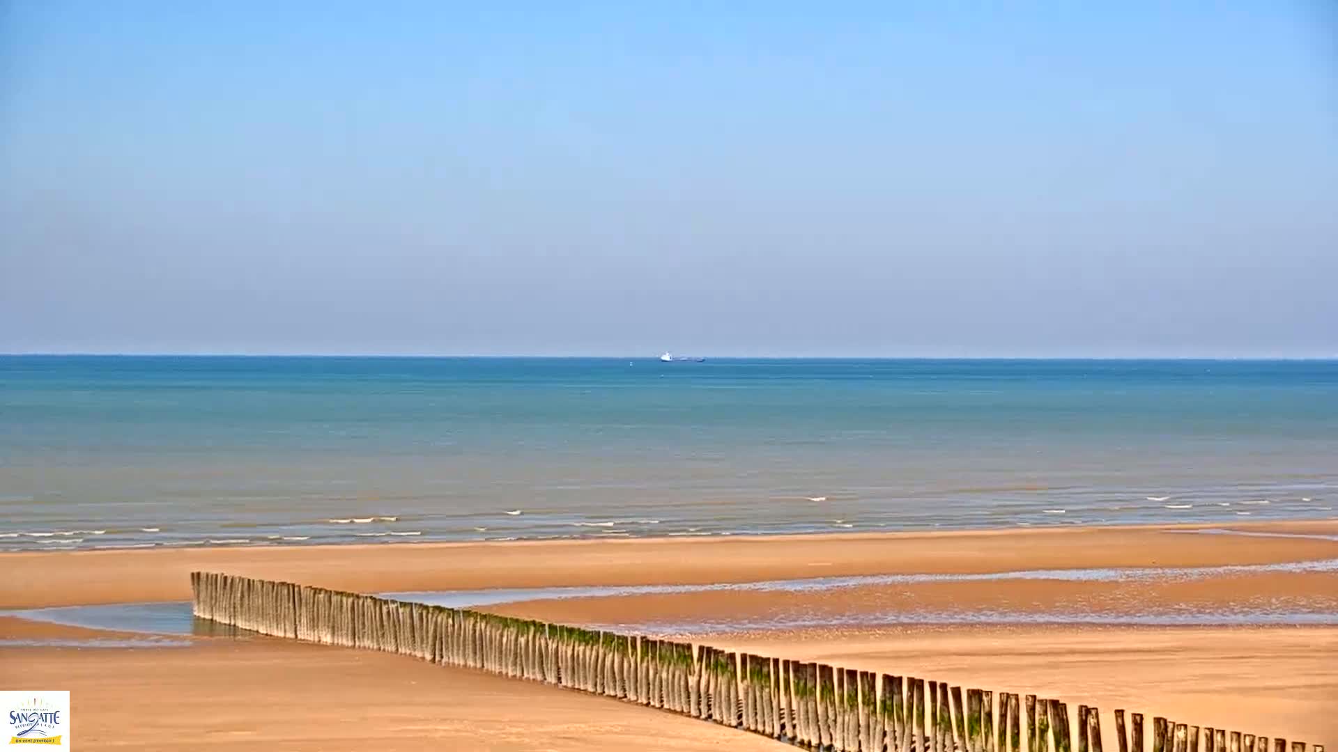 A sandy beach with a long, low wooden barrier extends into a calm, light-blue ocean under a clear sky.