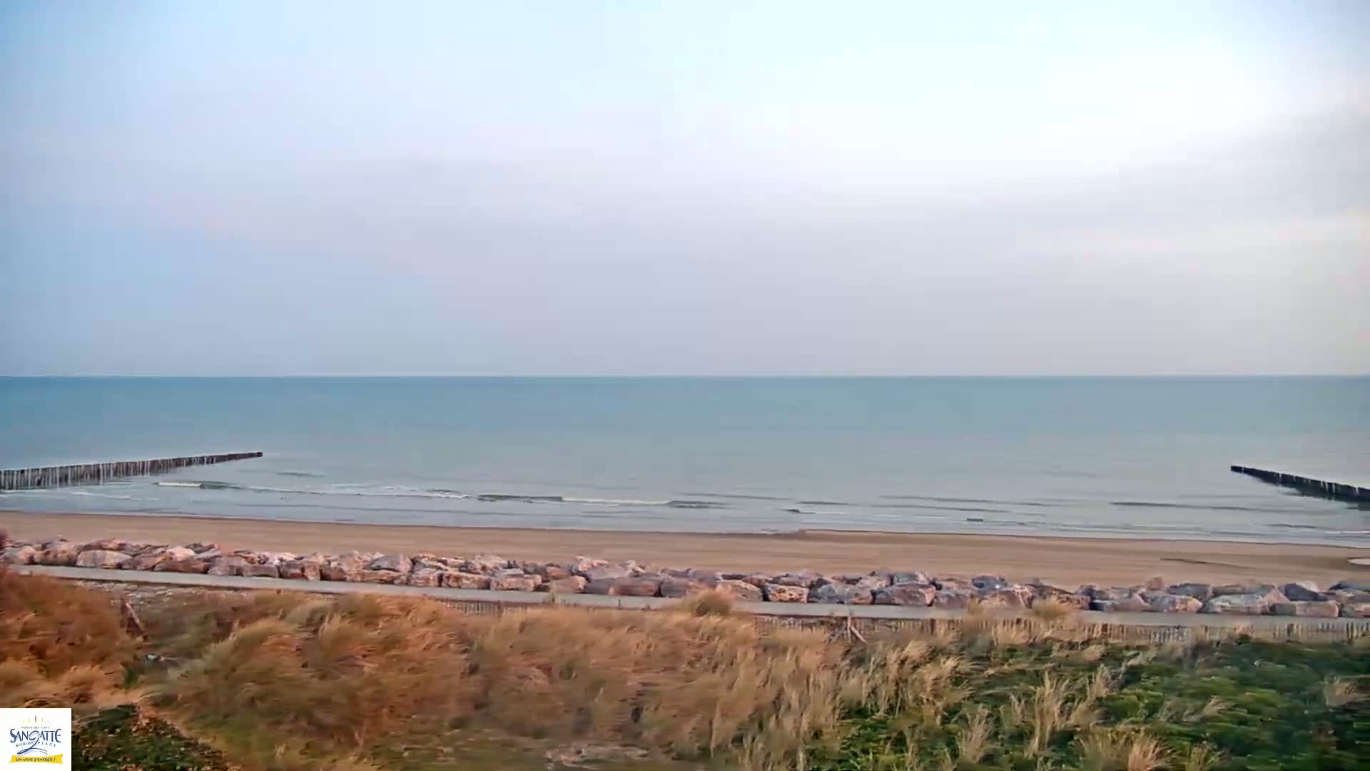 A sandy beach with calm ocean waves, two wooden breakwaters, and a line of rocks separating the beach from the dunes under a pale, clear sky.