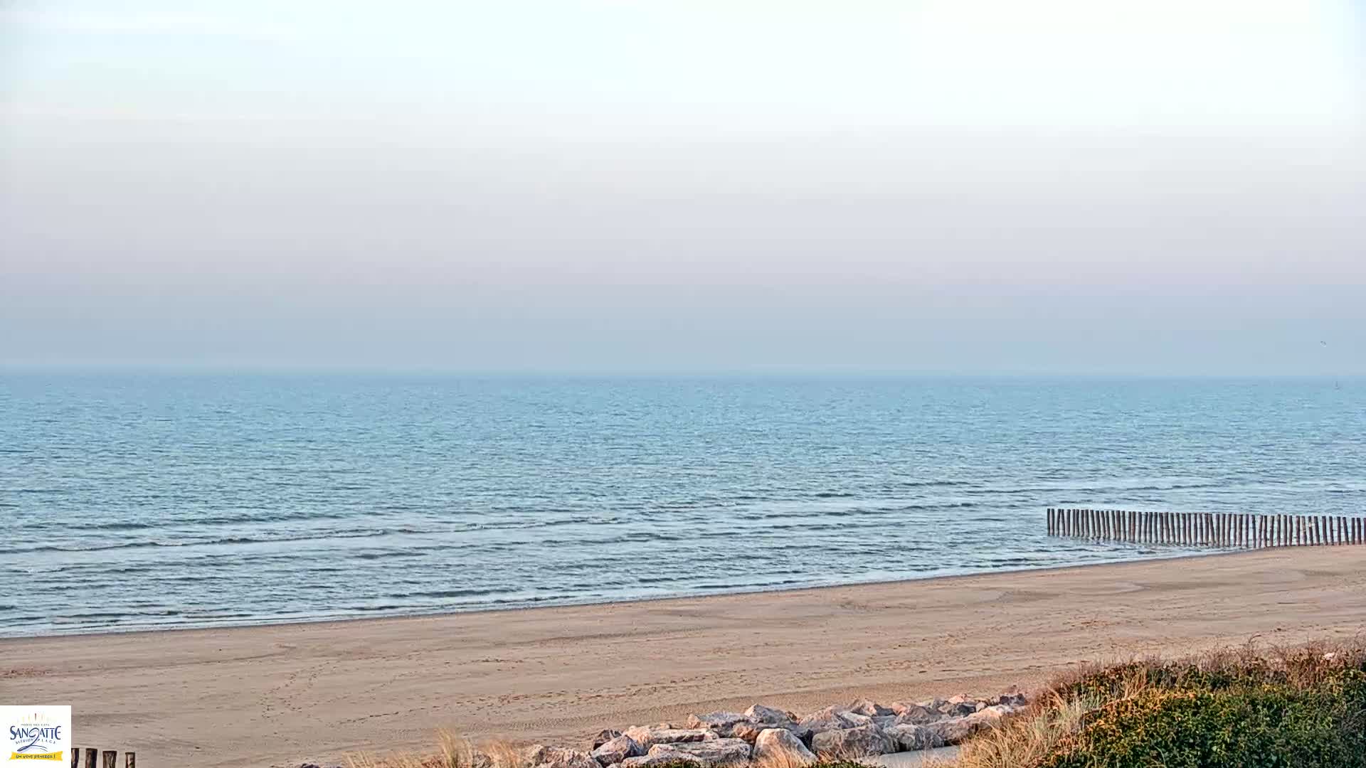 A sandy beach meets a calm, light blue ocean under a hazy sky.
