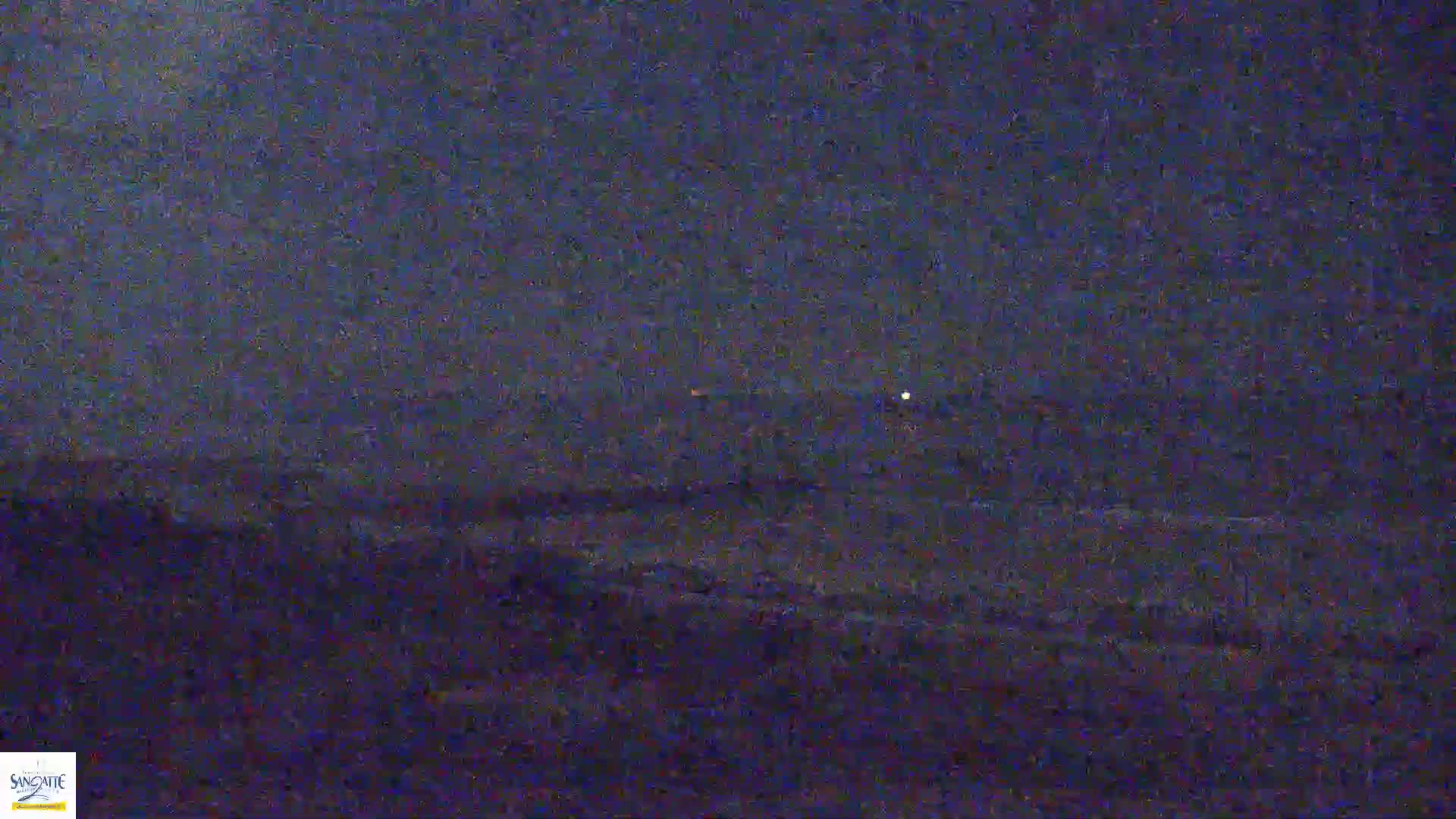 A dark, nighttime view of a wavy ocean under a cloudy sky with two distant lights visible on the horizon.