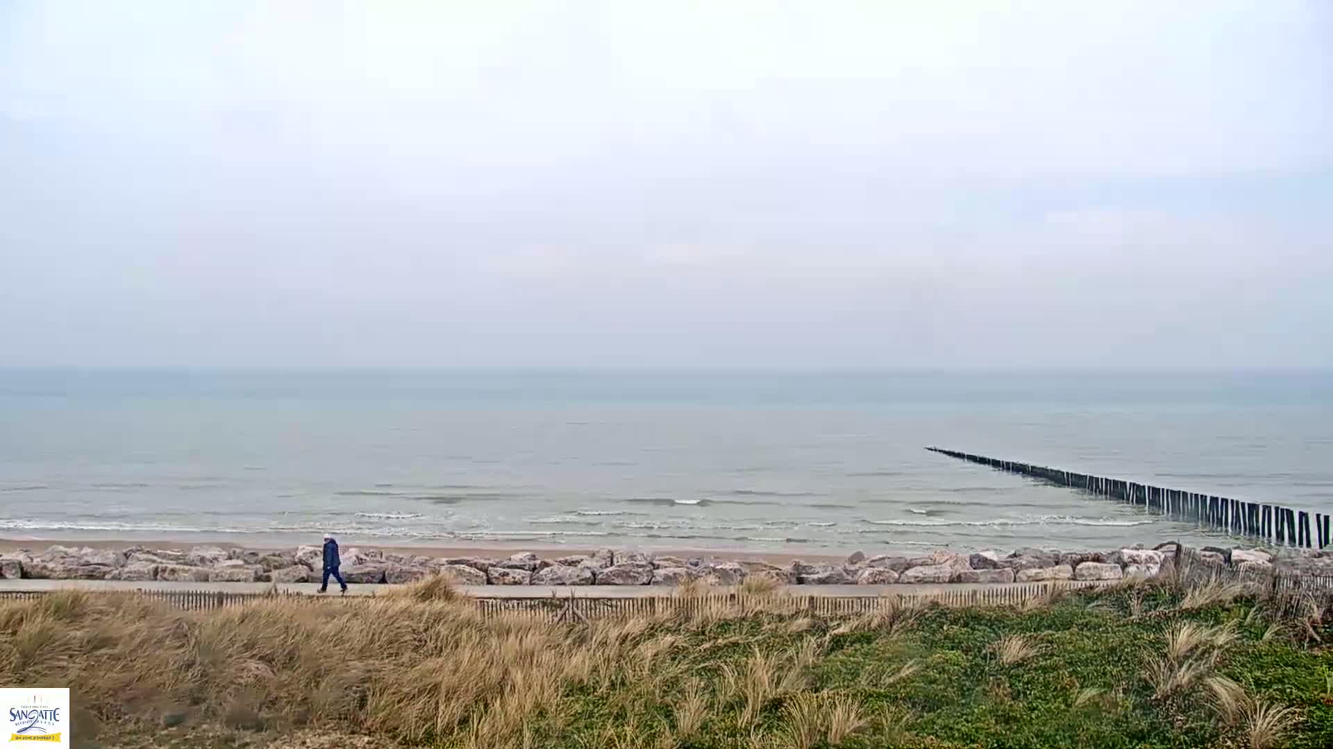 A person walks along a paved path overlooking a calm, gray sea with a long, thin breakwater extending from the shore on an overcast day.
