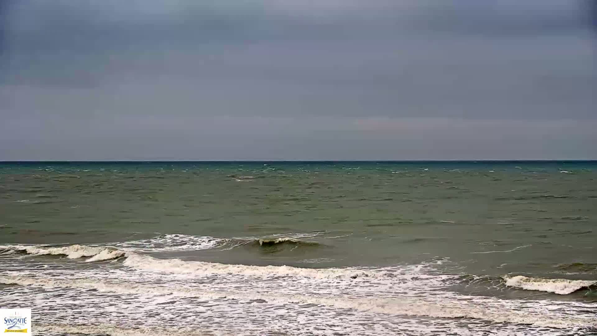 Under a gray sky, small waves break on a shore of a dark-green ocean.
