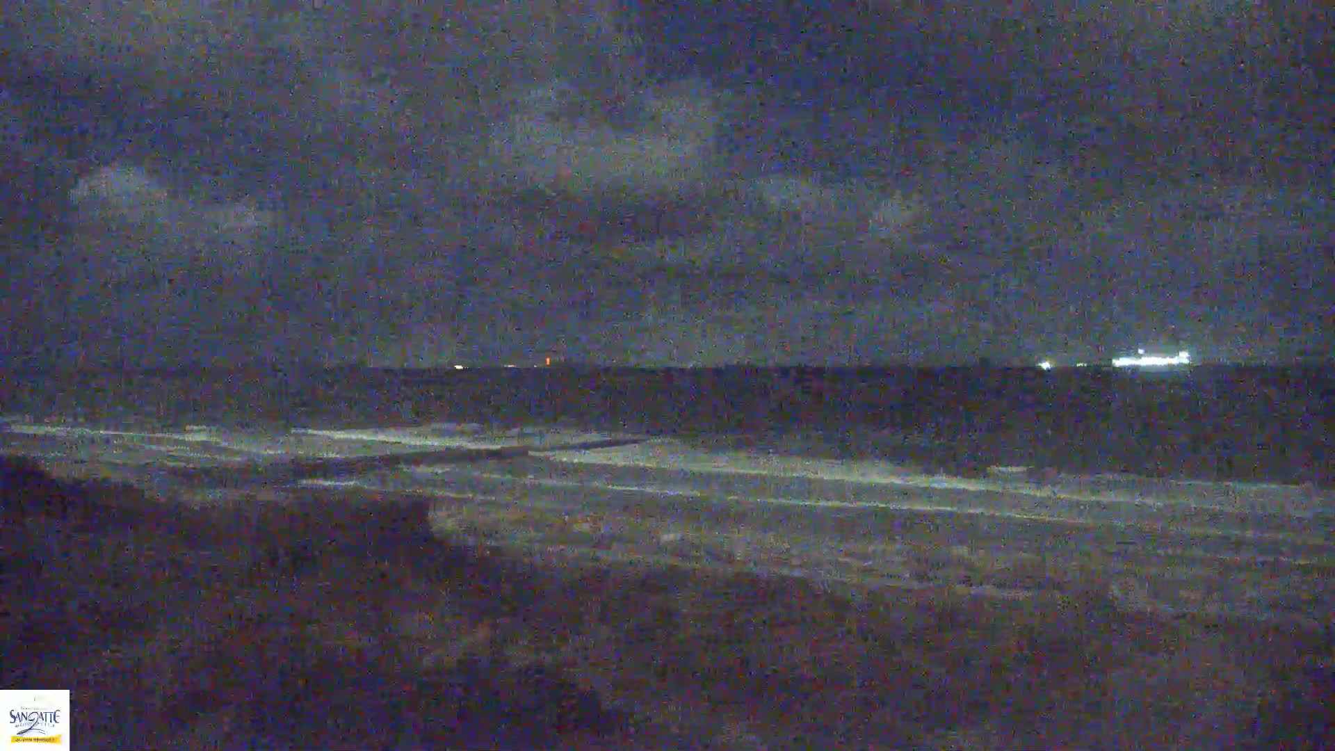 A dark, nighttime beach scene shows gently lapping waves on a shoreline under a cloudy sky.