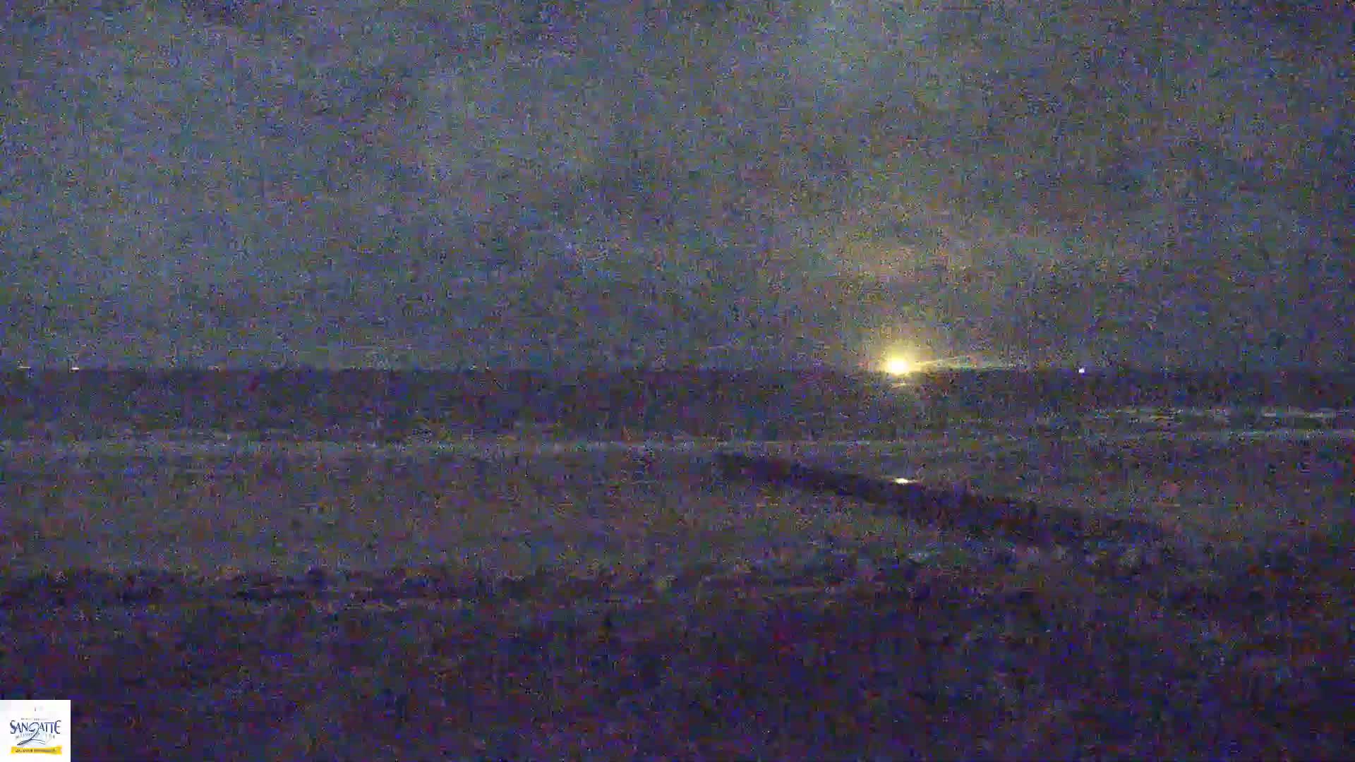 A dark, nighttime scene shows a body of water with a bright light on the horizon under a cloudy sky.