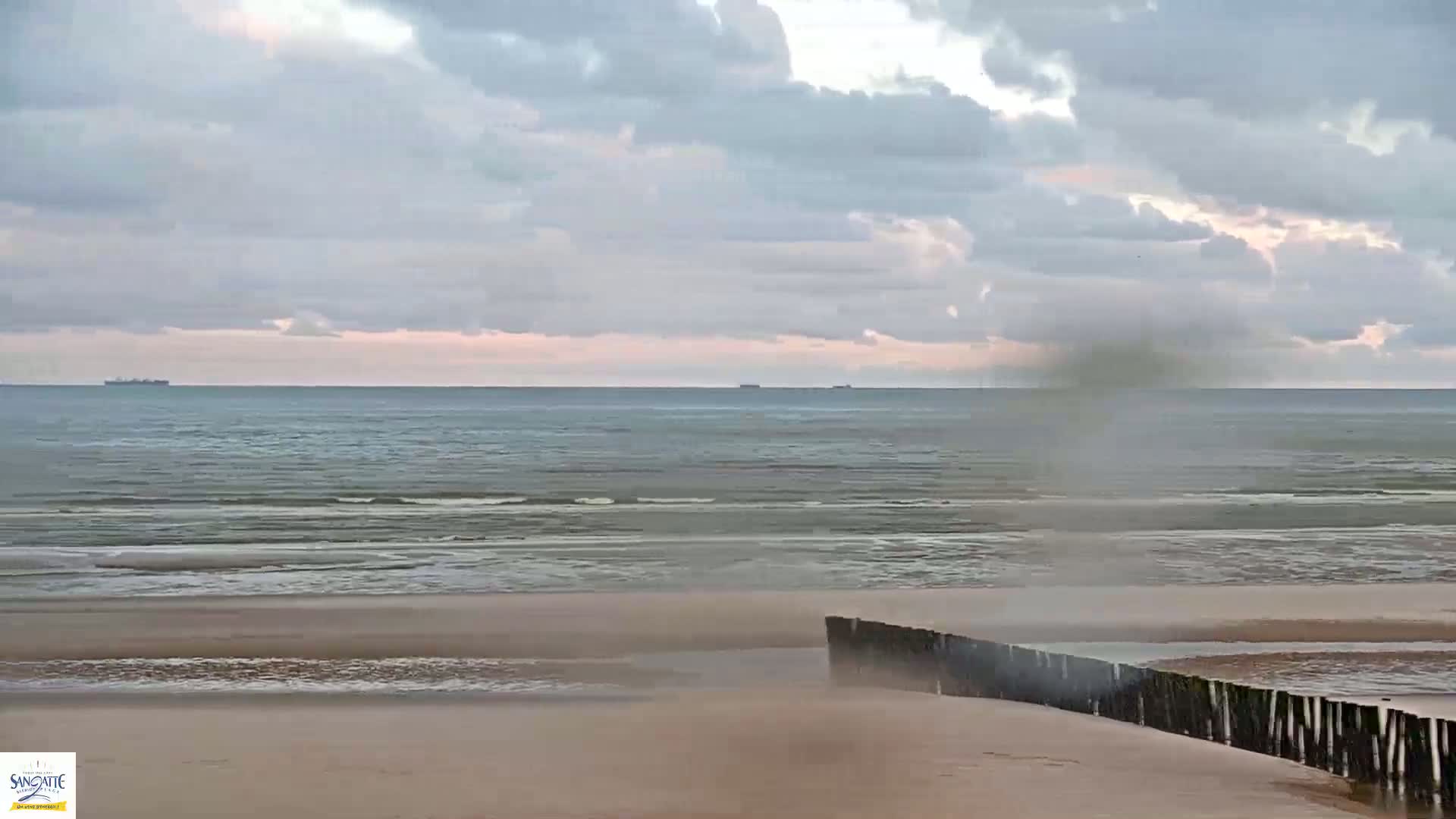 A sandy beach with a low wooden barrier extends into a calm sea under a cloudy sky, with a small plume of spray or mist visible near the barrier.