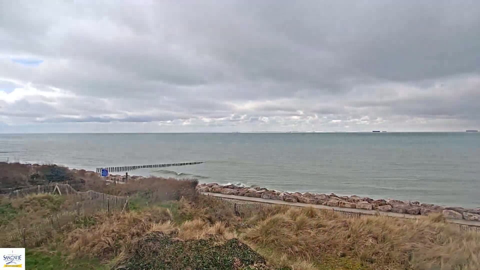 A partly cloudy sky overlooks a gray sea with small waves breaking against a rocky shore, partially protected by a wooden breakwater.