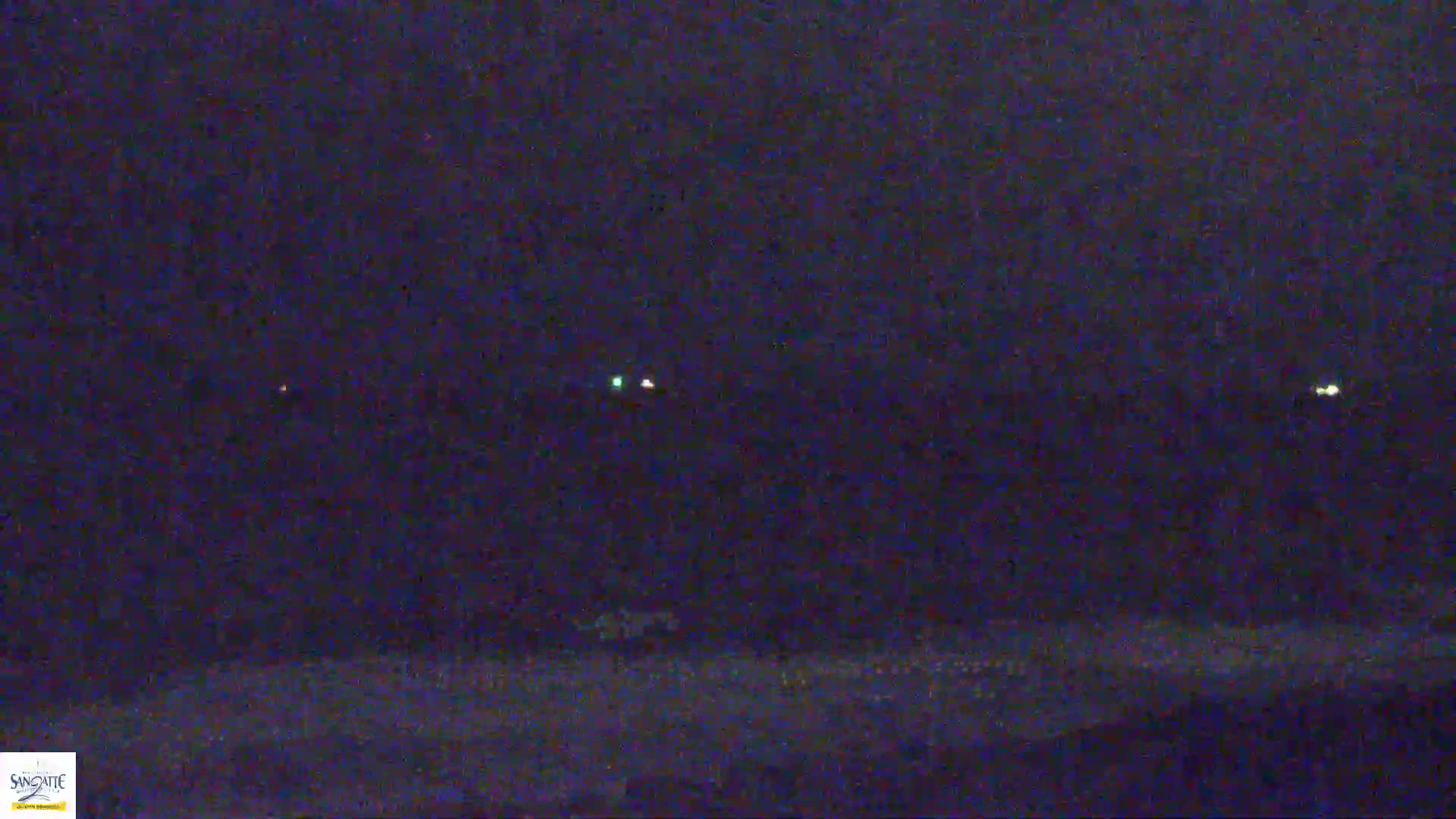 A dark, nighttime scene shows a flat, dark landscape with several small, faint lights visible in the distance.