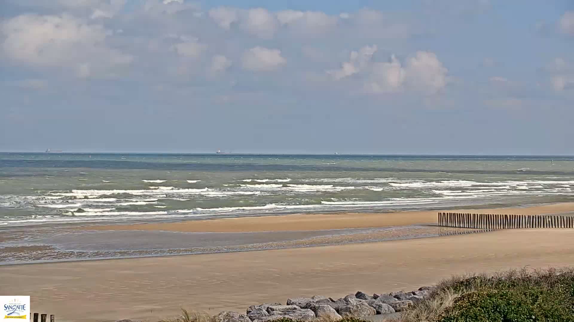 A sandy beach with a moderately windy sea under a partly cloudy sky features a line of wooden posts separating the beach from the water.