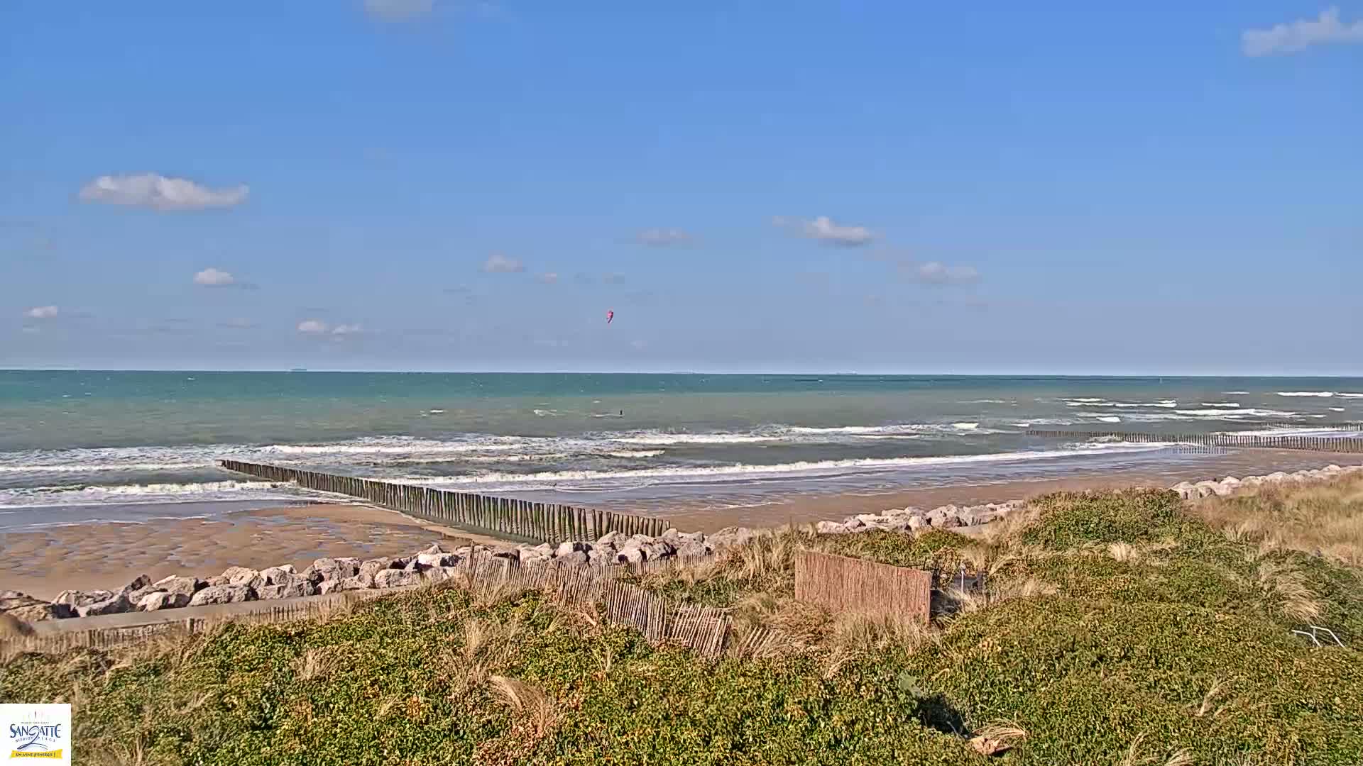 A sandy beach with moderate waves and a kitesurfer is visible under a mostly sunny sky.
