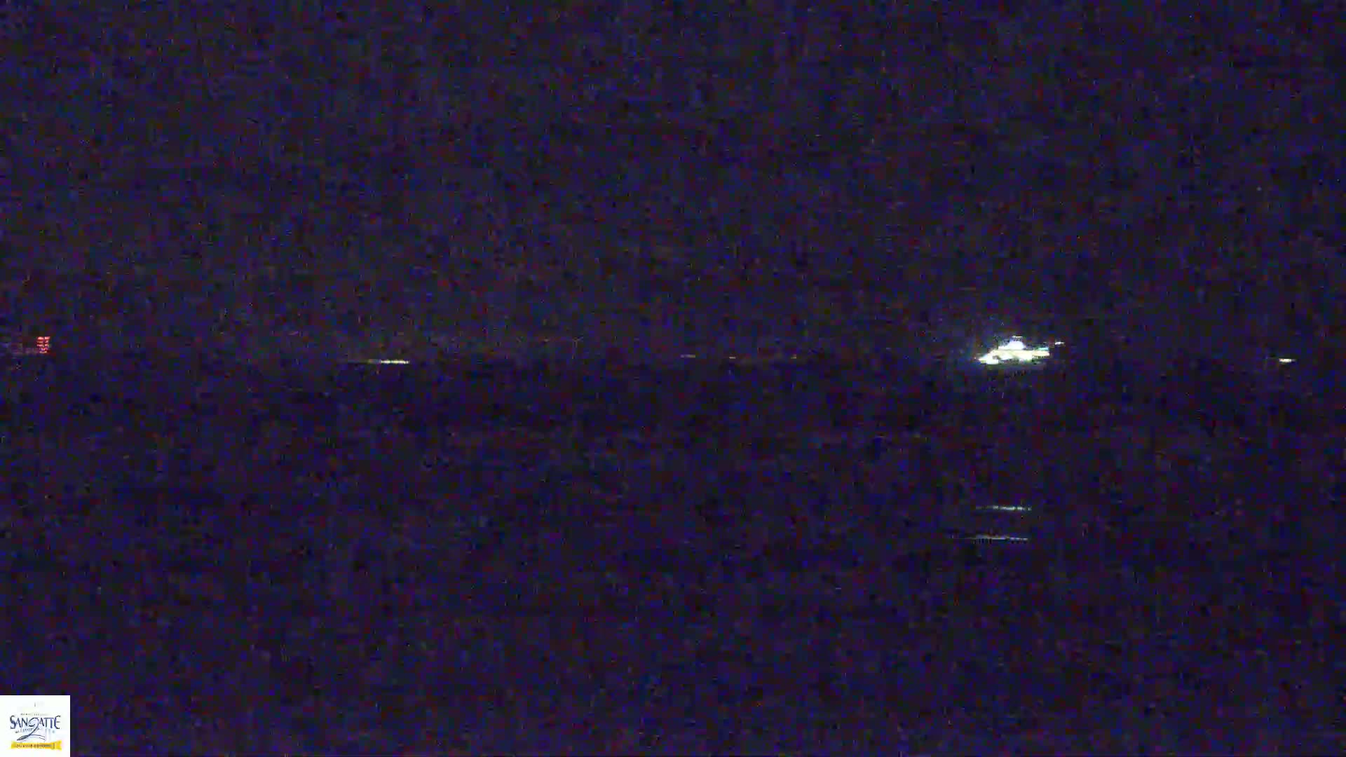 A dimly lit nighttime scene shows a body of water with faint lights on the horizon under a dark sky.
