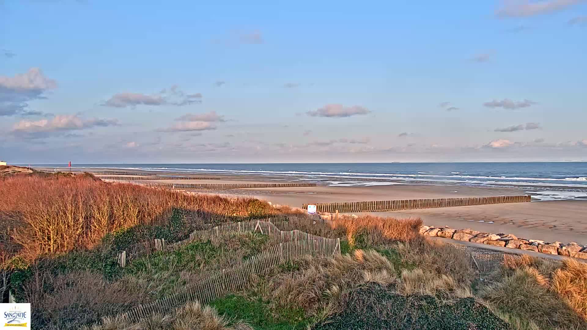 A sandy beach with calm waves under a partly cloudy sky is partially protected by wooden barriers and bordered by low-lying vegetation.