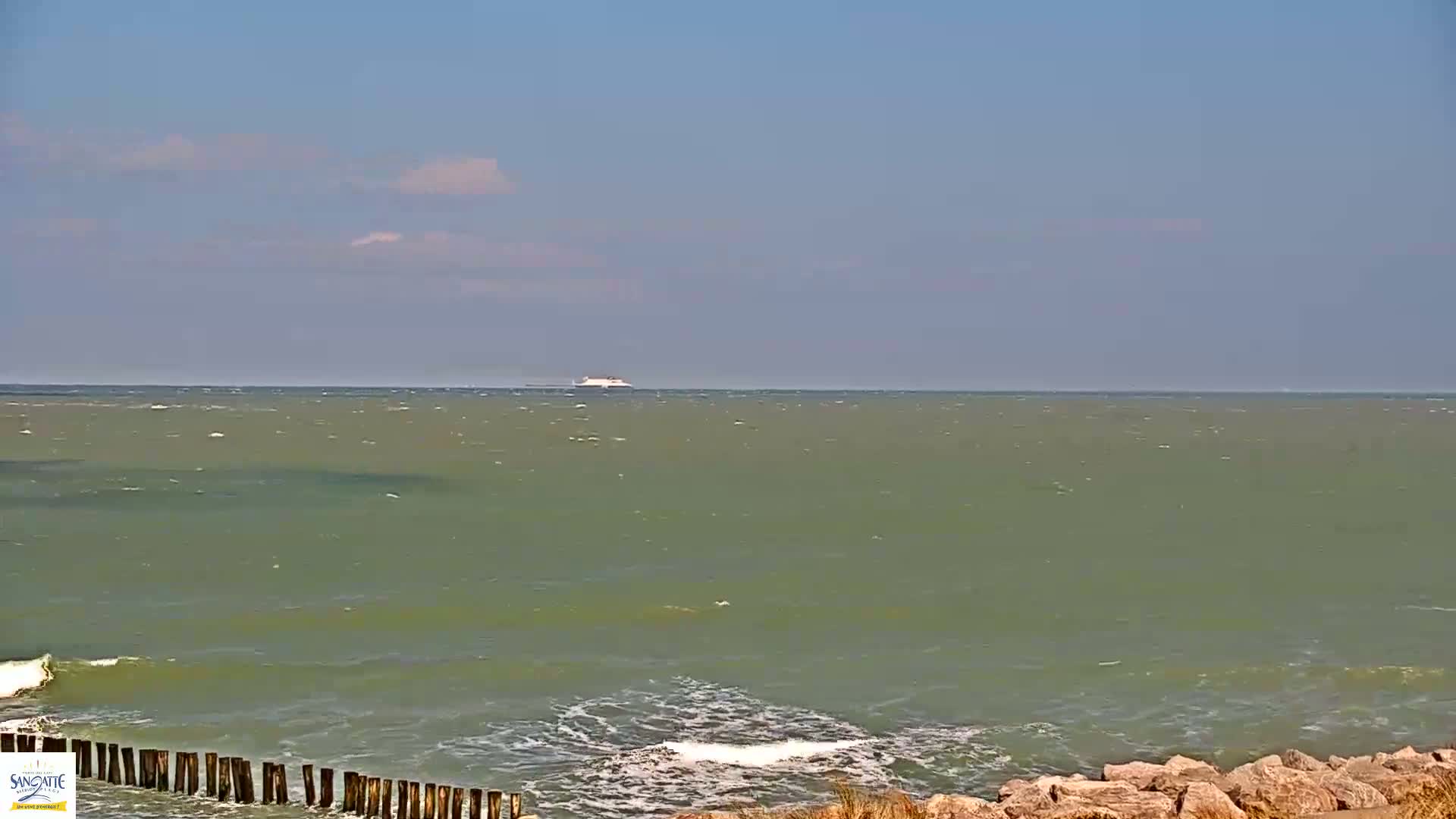 A large body of somewhat choppy, greenish-blue water stretches to the horizon under a mostly clear, light blue sky, with a distant ship visible on the water.