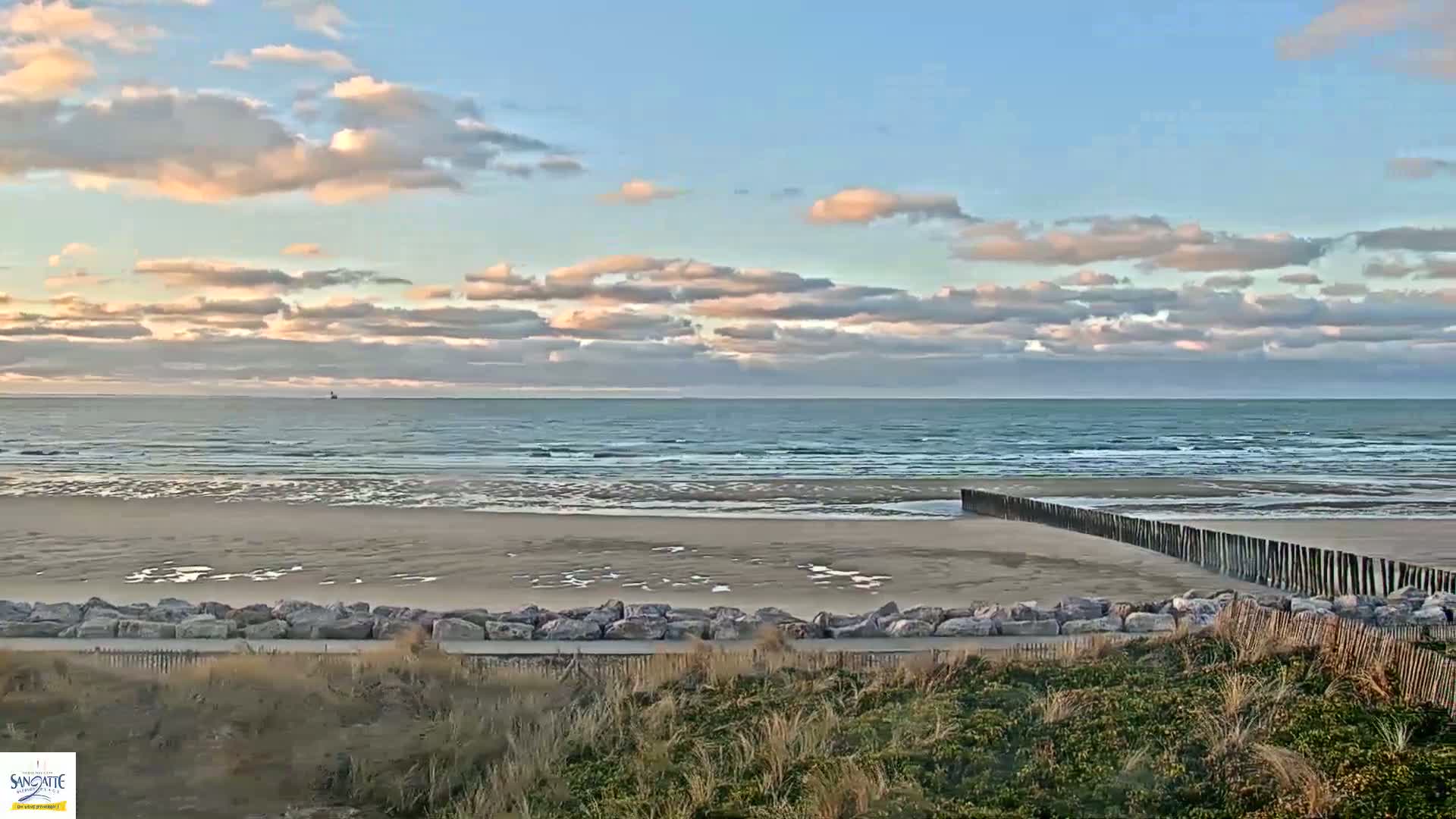 A sandy beach with a calm sea under a partly cloudy sky is viewed from a dune overlooking a rock wall.