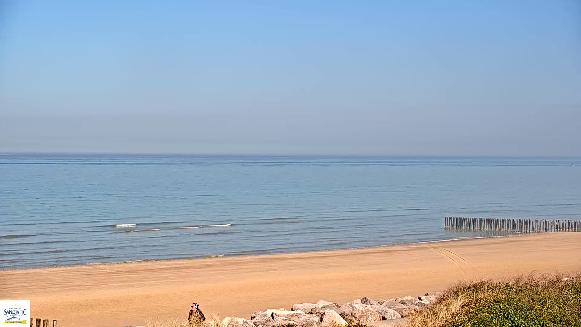 A sandy beach with calm, light blue water and a clear sky stretches to the horizon, featuring a line of wooden posts in the water near the shore and a lone person standing near the dune line.
