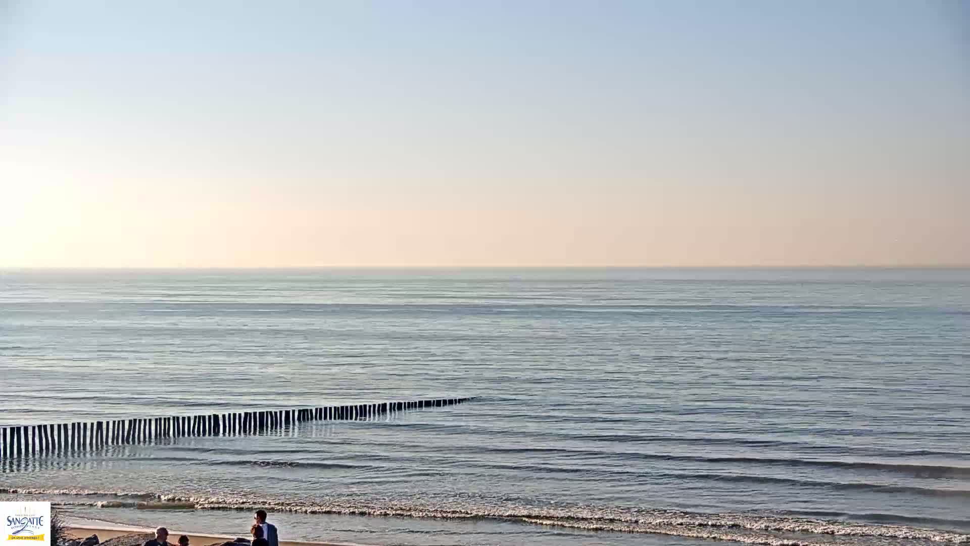 A calm, pale-blue sea stretches to the horizon under a hazy, light-pink sky, with a line of wooden posts partially submerged in the shallows and a few people on the beach.