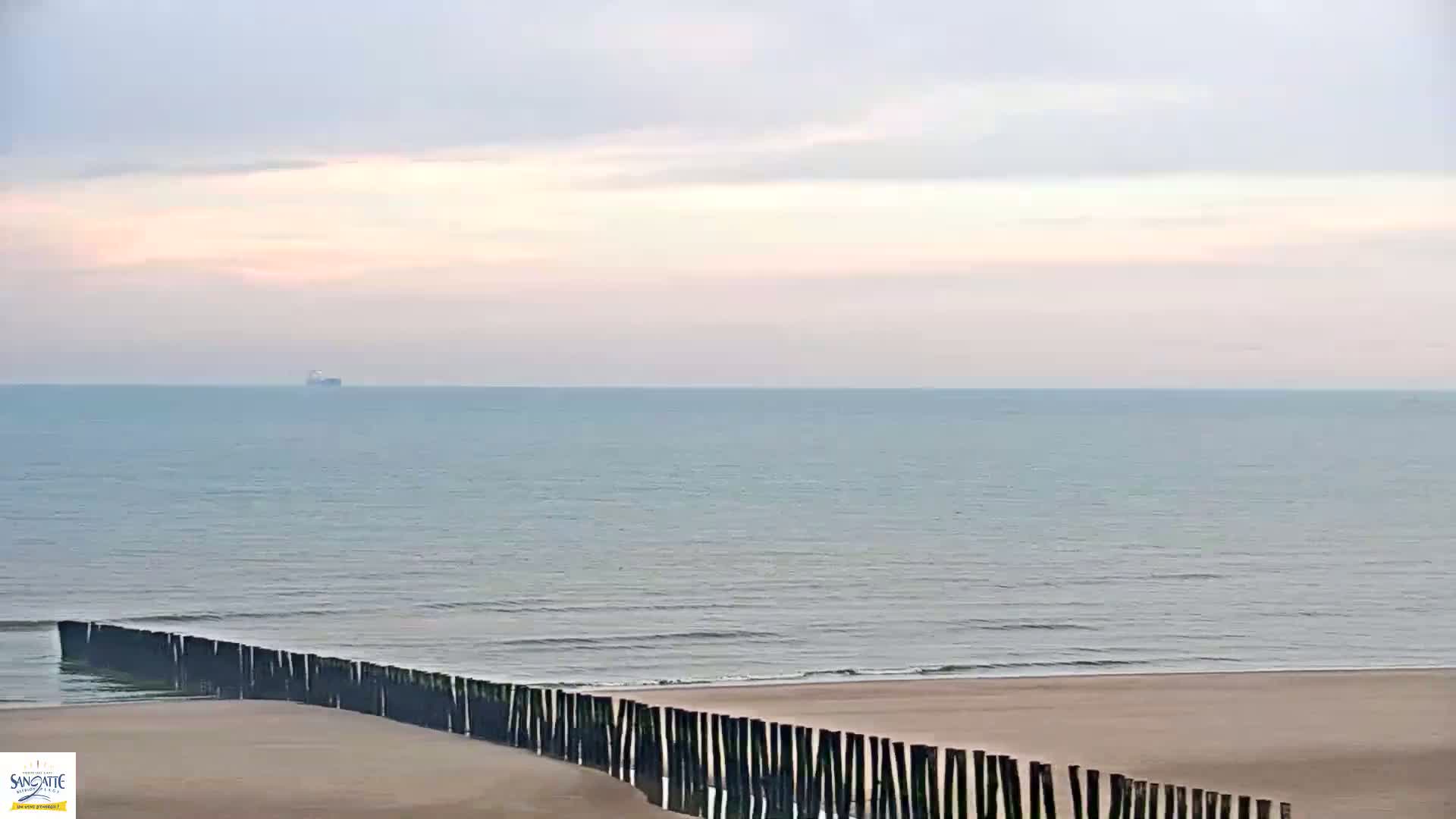 A calm sea with a distant ship under a pale sky meets a sandy beach protected by a long, dark wooden barrier.