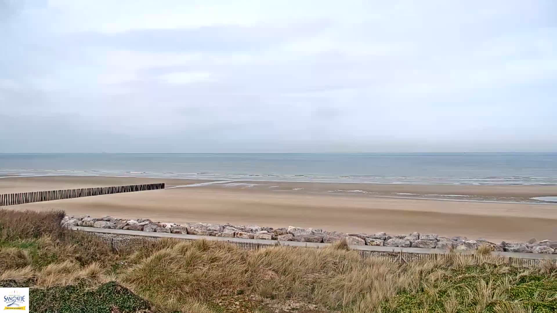 A wide sandy beach stretches to a calm, grey sea under a cloudy sky, protected by a low wooden barrier.