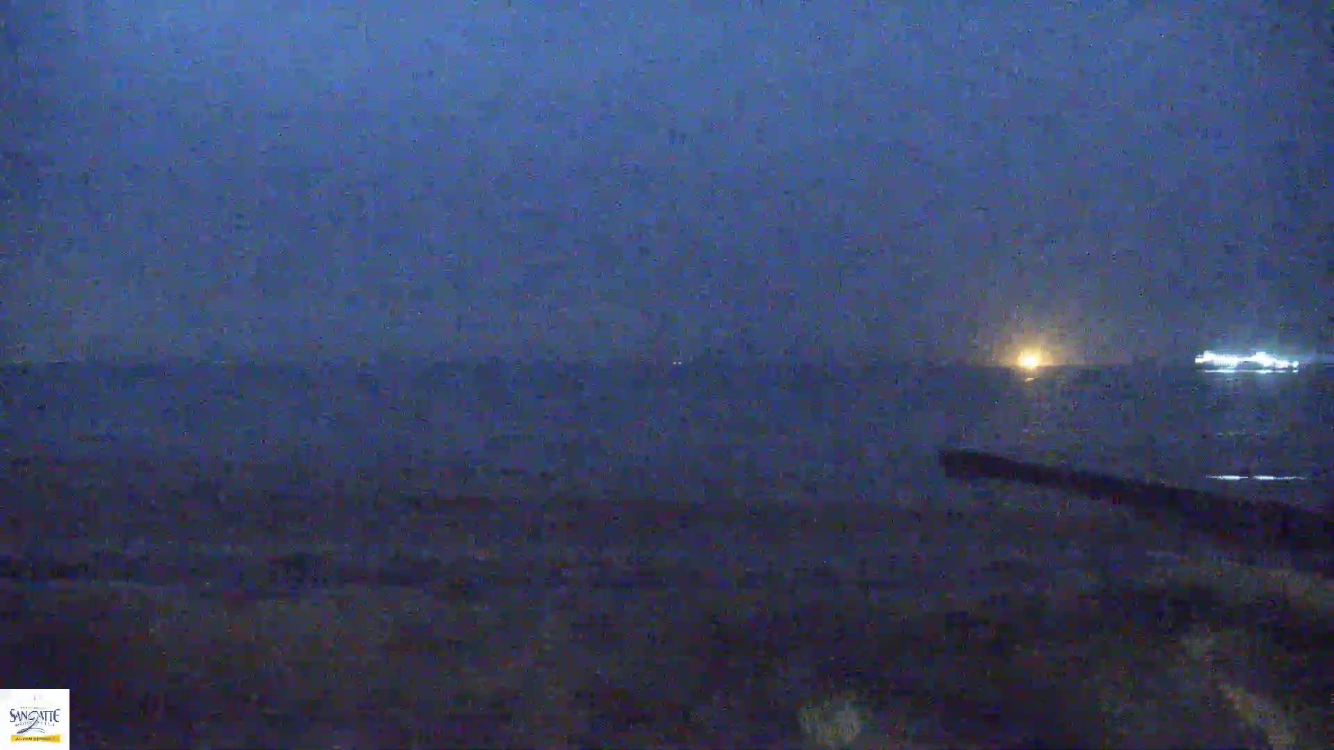 A dark, nighttime scene shows a body of water with faint lights on the horizon under a mostly clear, dark blue sky.