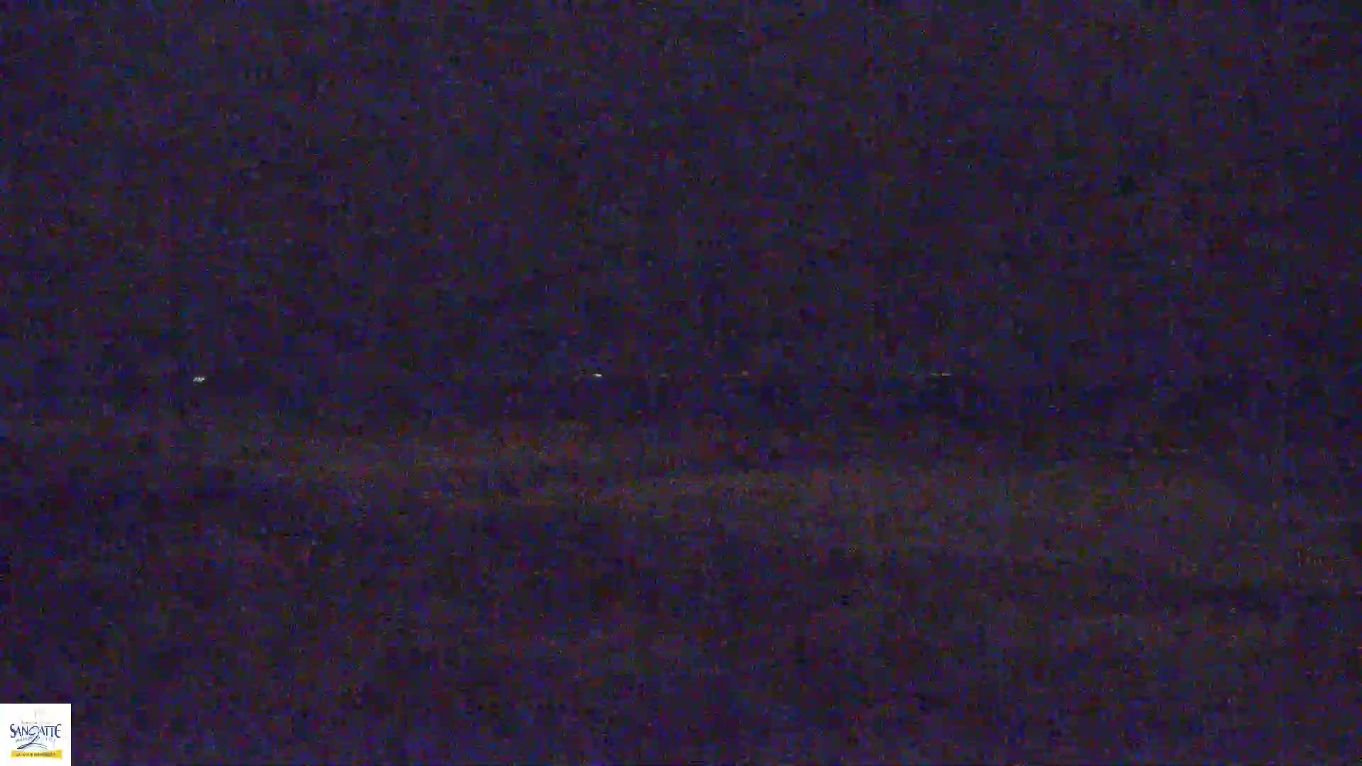 A dark, nighttime scene shows a distant line of faint lights across a blurry, indistinct landscape.