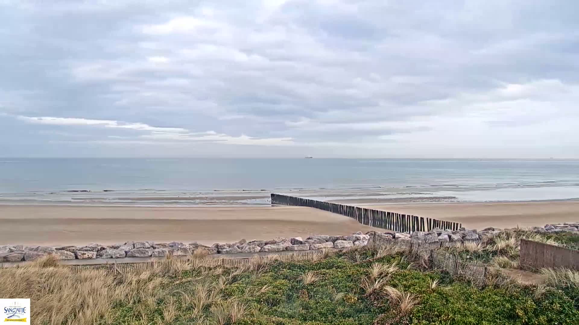 A sandy beach with a long, low wooden barrier extending into the calm, light-blue ocean under a cloudy sky.