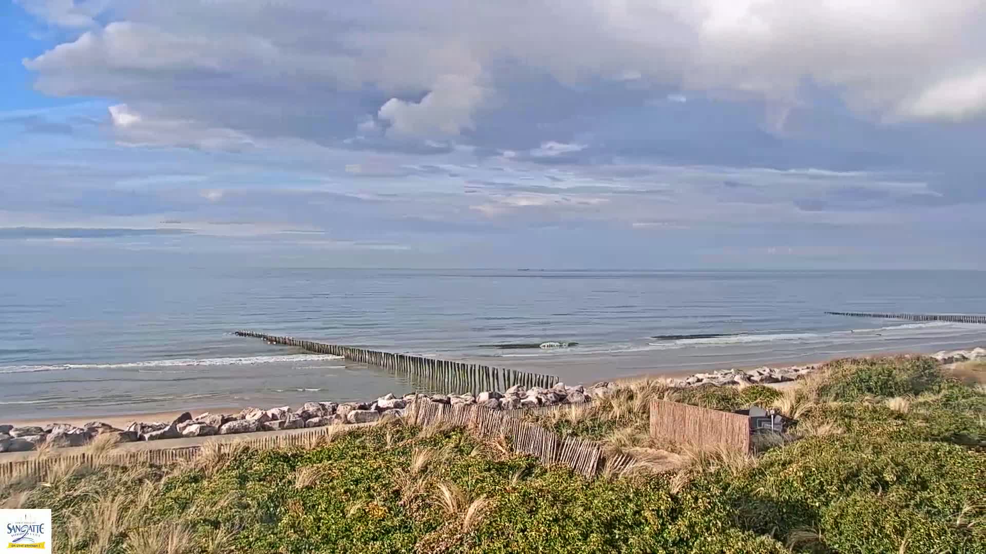 A partly cloudy day shows a calm ocean with a sandy beach and rock barriers, viewed from a dune covered in low vegetation.