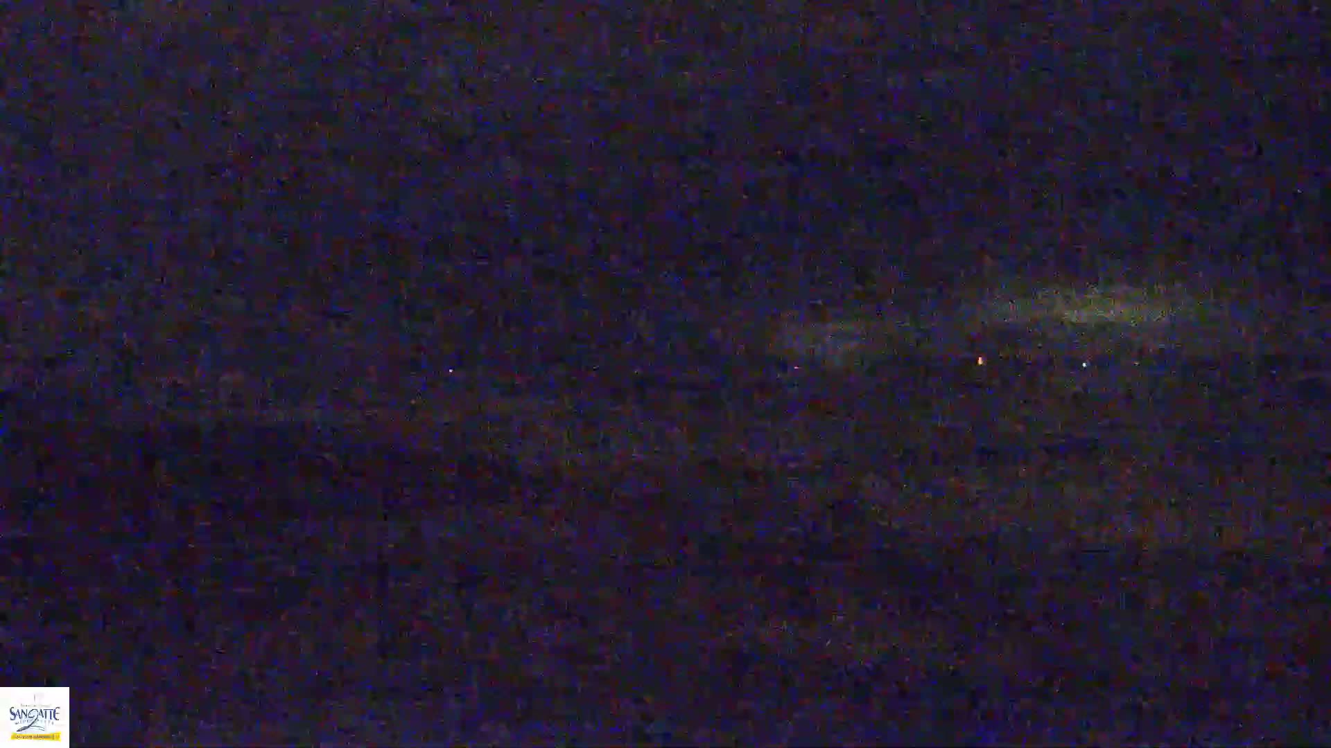 A nighttime, low-light image shows a dark, possibly cloudy, sky with a few faint, distant lights on the horizon.
