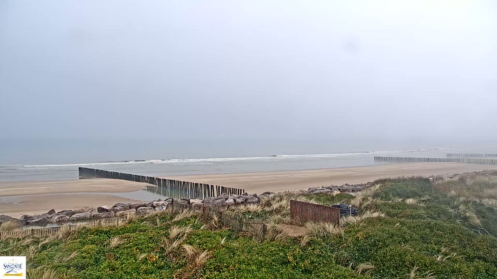 A foggy beach scene shows a sandy shore with a low wooden barrier partially submerged in the water, separated from the dunes by a line of rocks.