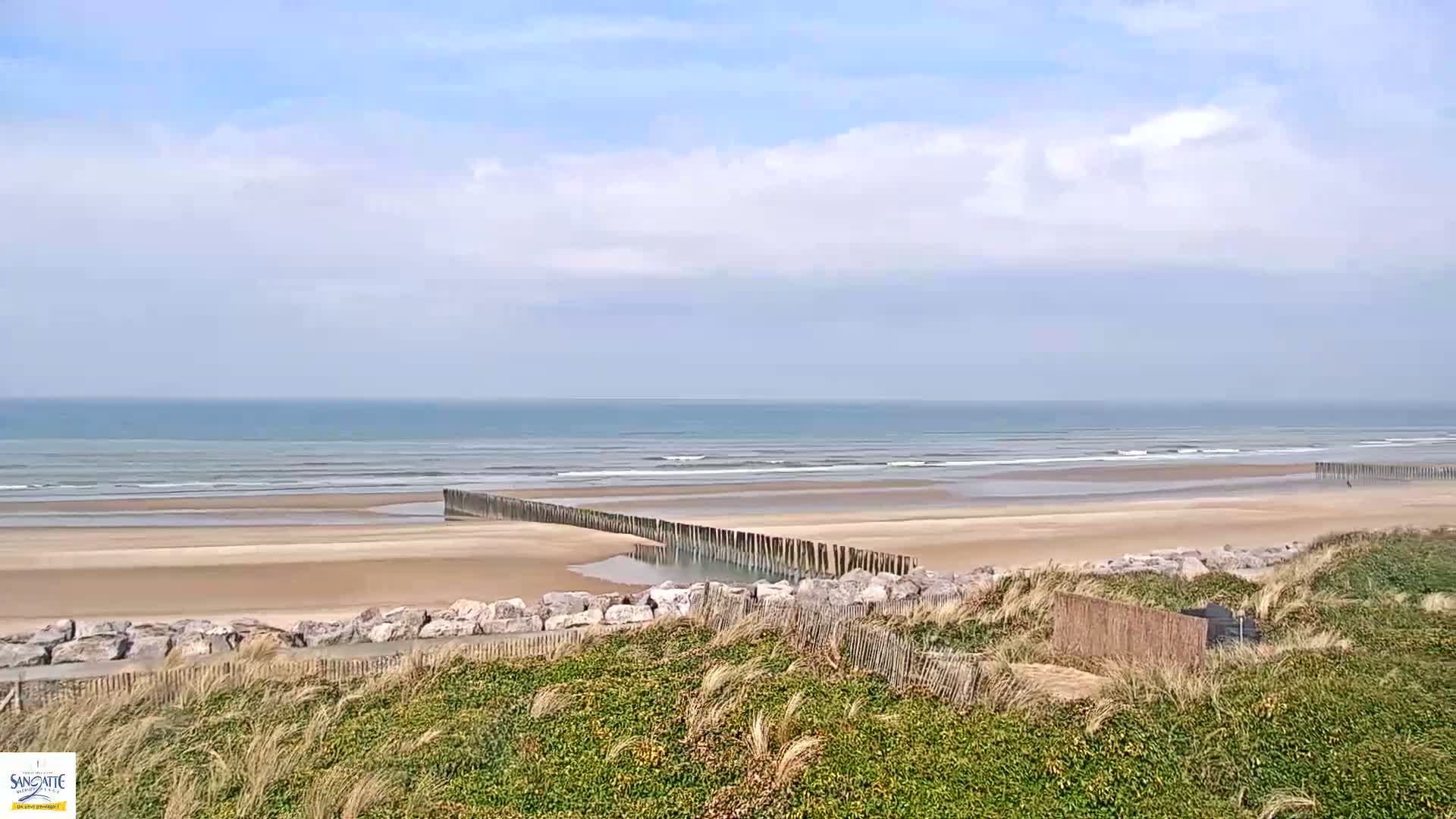 A sandy beach with a calm ocean and a partially submerged wooden barrier stretches out under a partly cloudy sky.