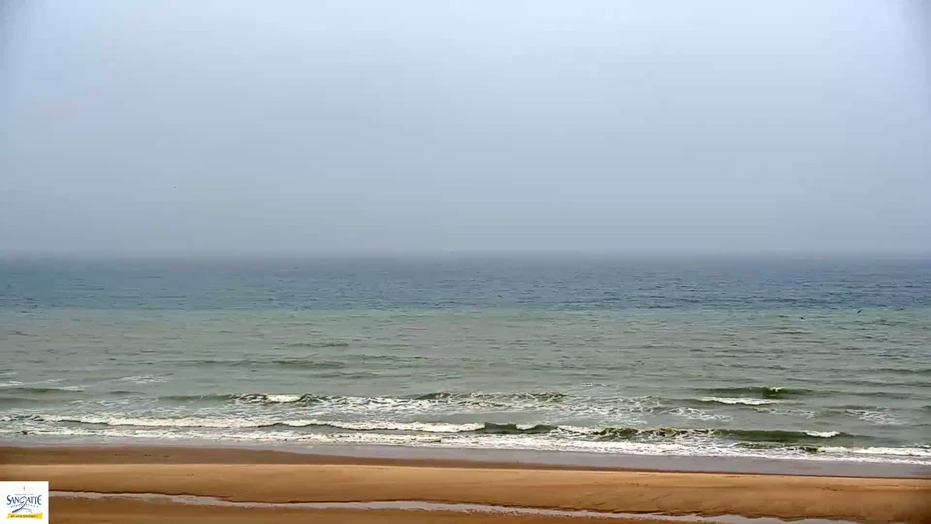 A foggy day at the beach shows a calm sea with small waves lapping a sandy shore.