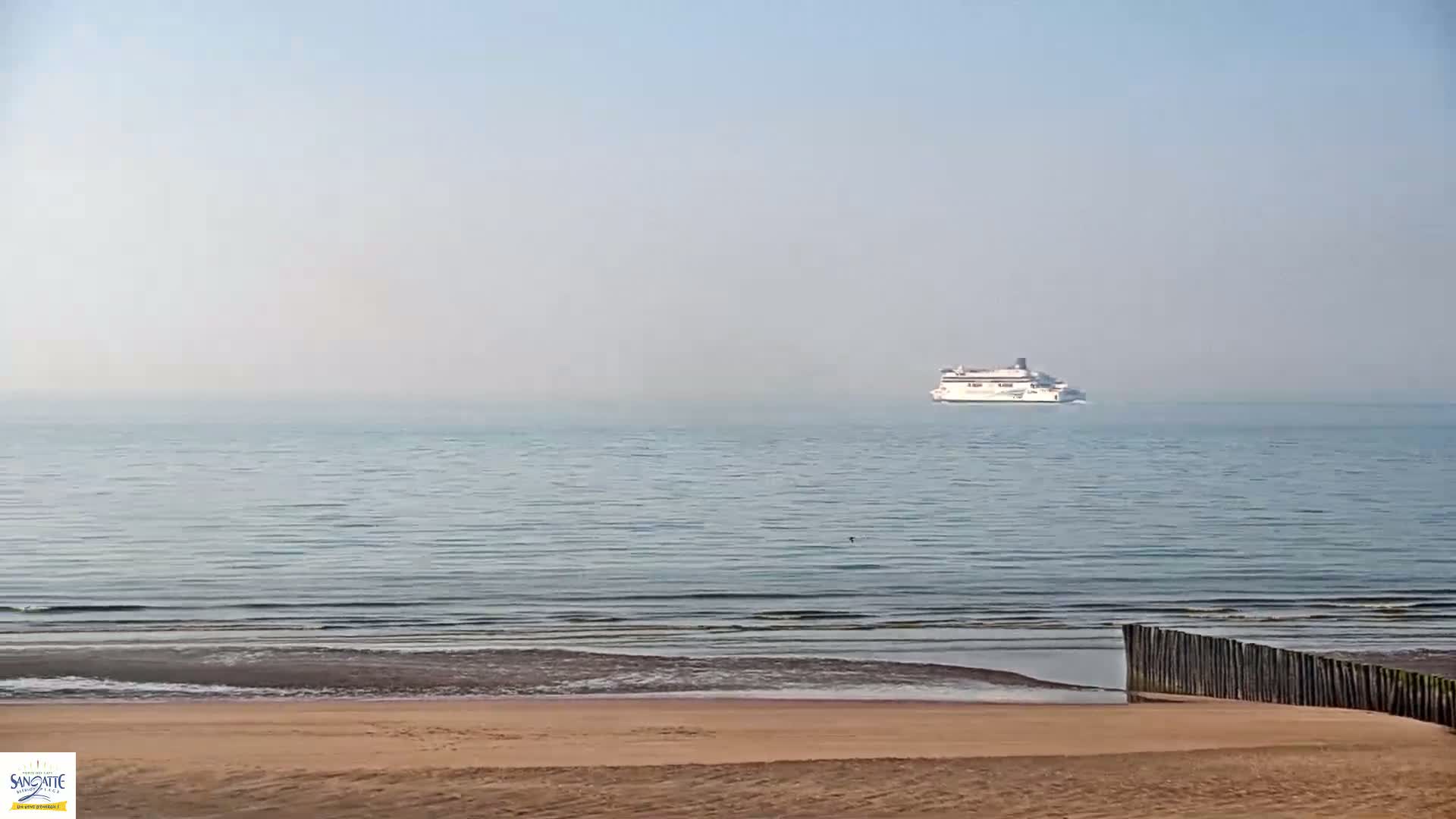 A hazy day shows a ferry on calm ocean waters beyond a sandy beach with a wooden barrier.