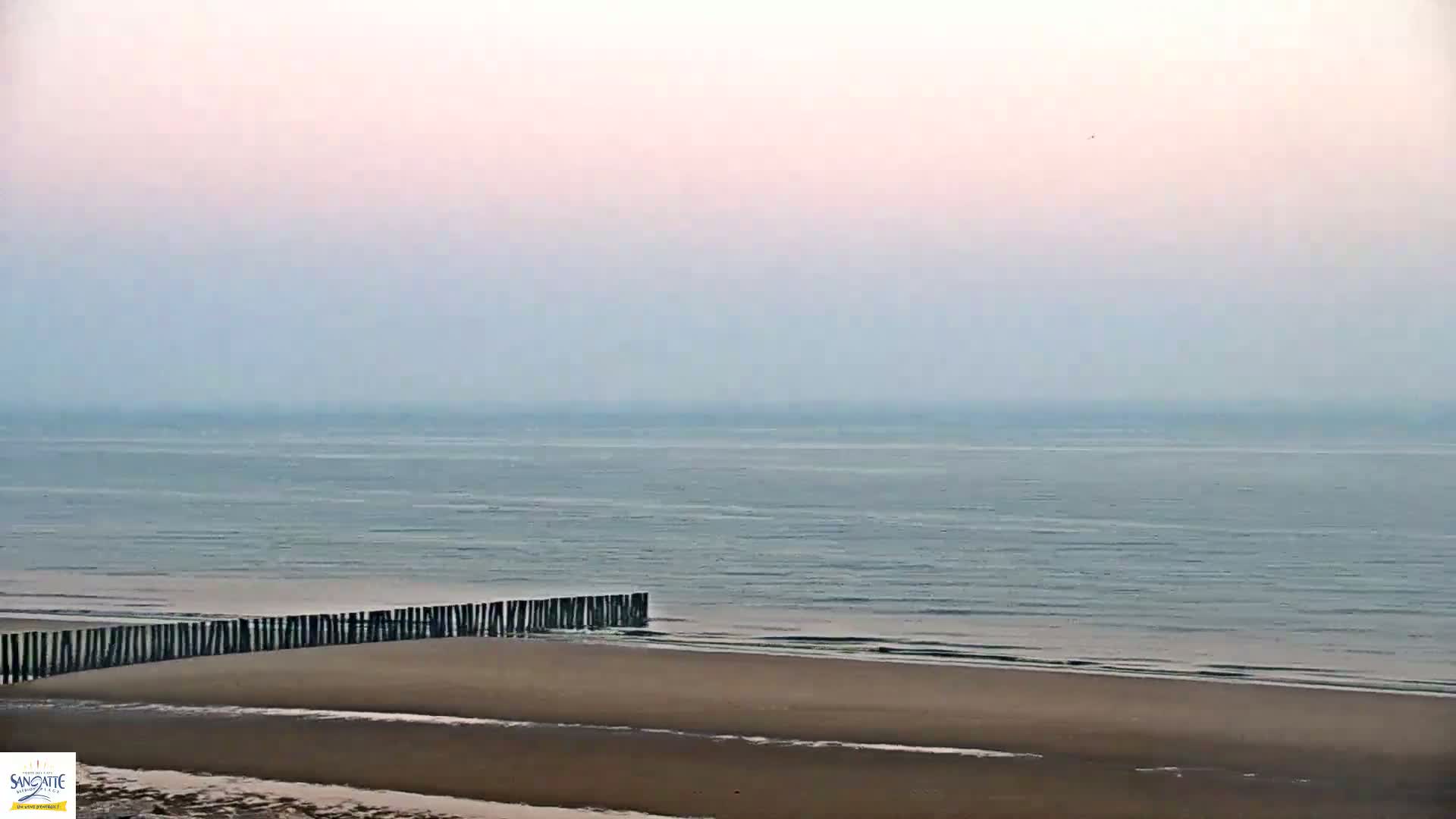 A hazy, pale-pink sky overlooks a calm sea meeting a sandy beach protected by a low, dark wooden barrier.