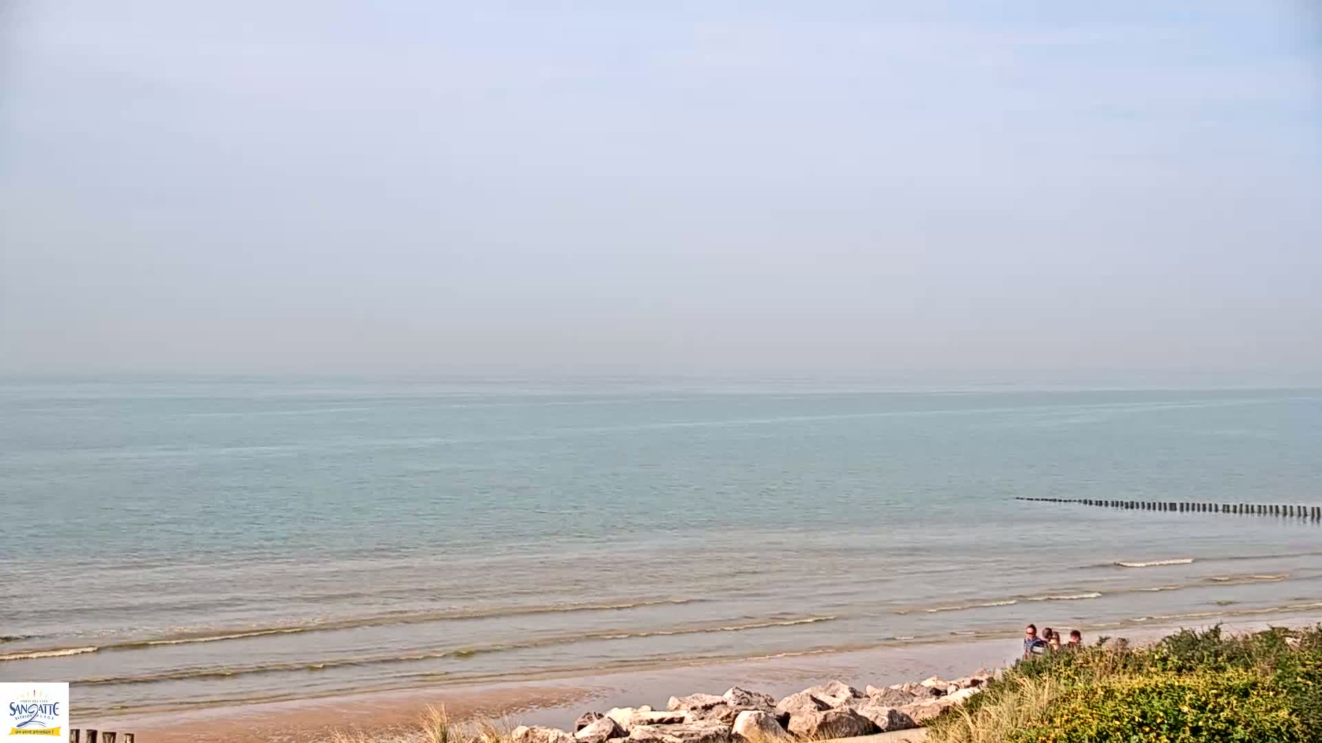 A hazy day at a beach shows calm, light teal water lapping gently on a sandy shore where a few people stand near some rocks and vegetation.