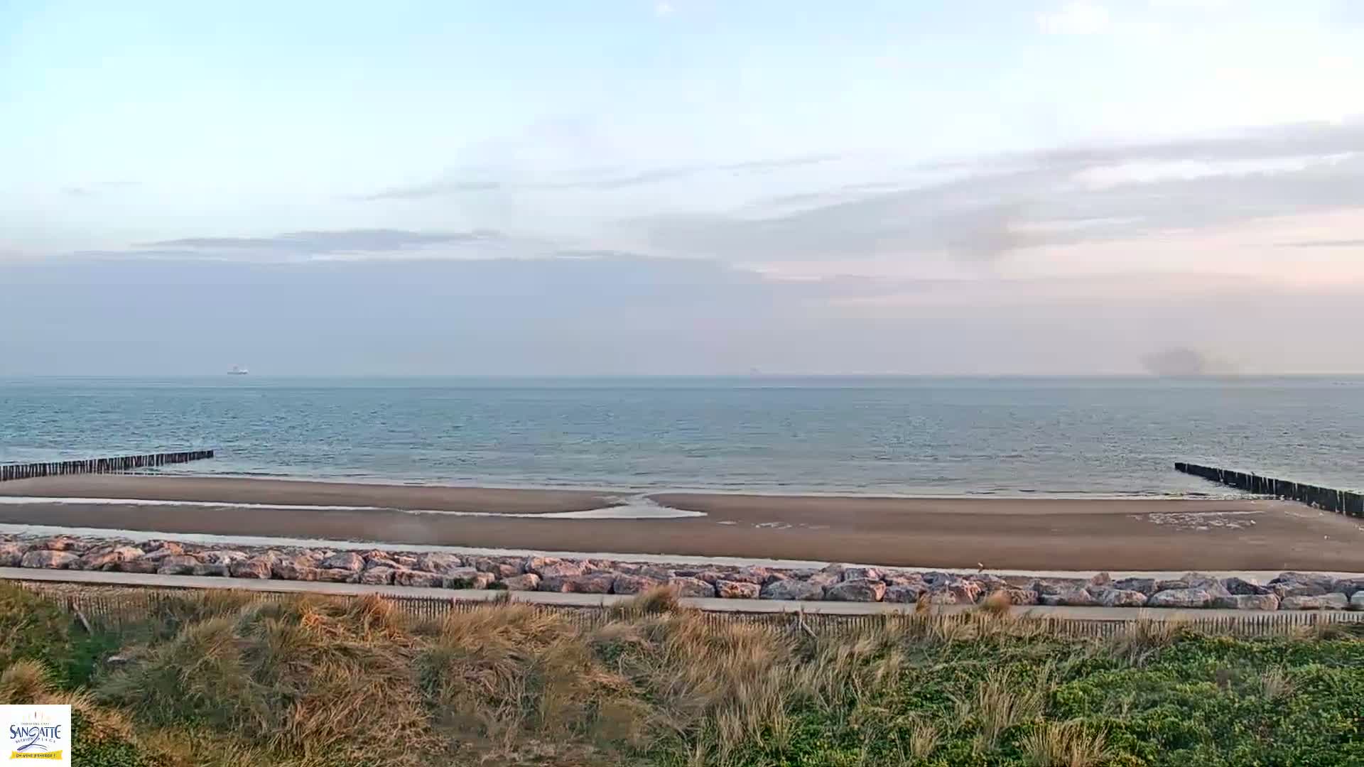 A calm ocean meets a sandy beach protected by rock barriers and dunes under a mostly cloudy sky.