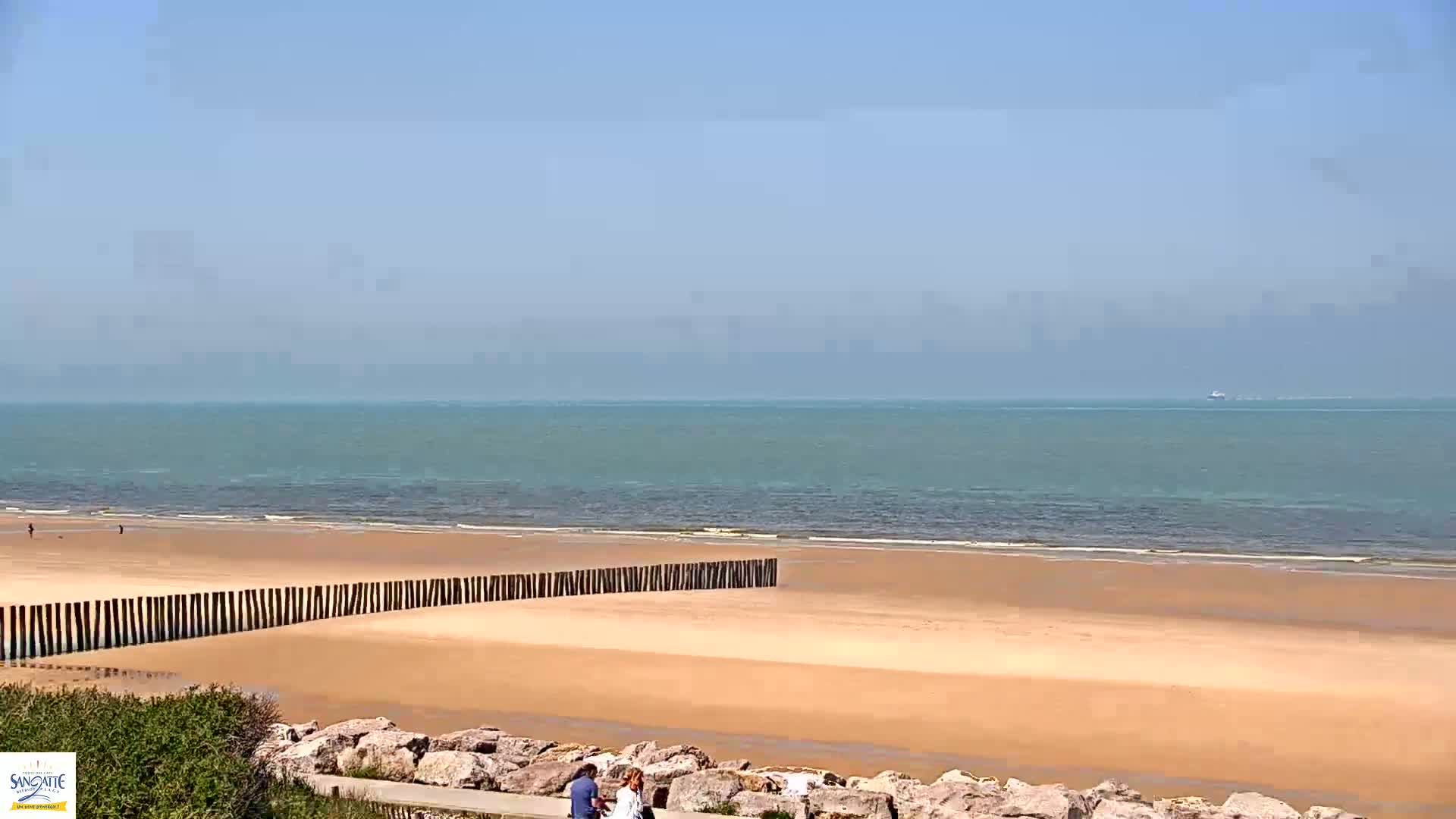 A sandy beach stretches to a calm, teal ocean under a clear, bright blue sky, with a few people walking near a low rock wall.