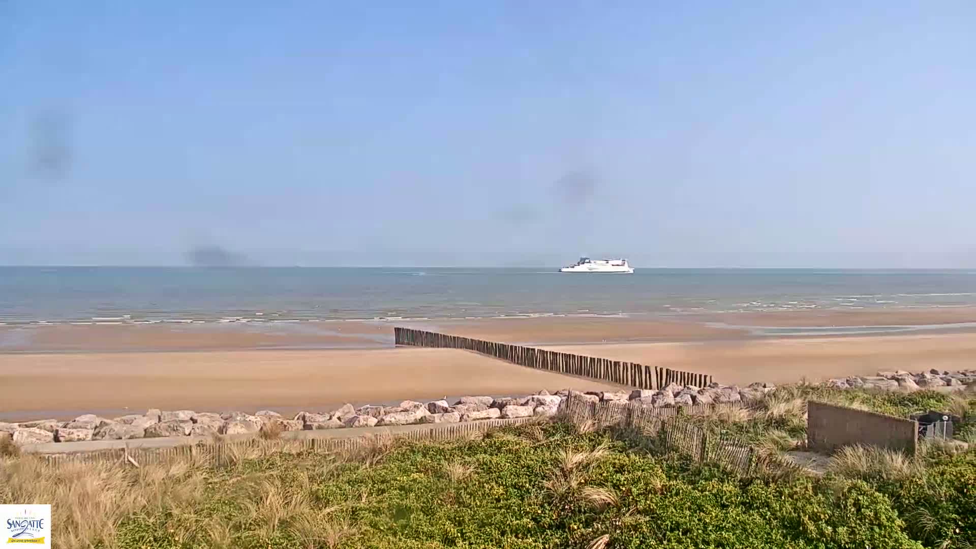 A sandy beach stretches out to a calm sea where a ferry is visible on the horizon under a clear blue sky.
