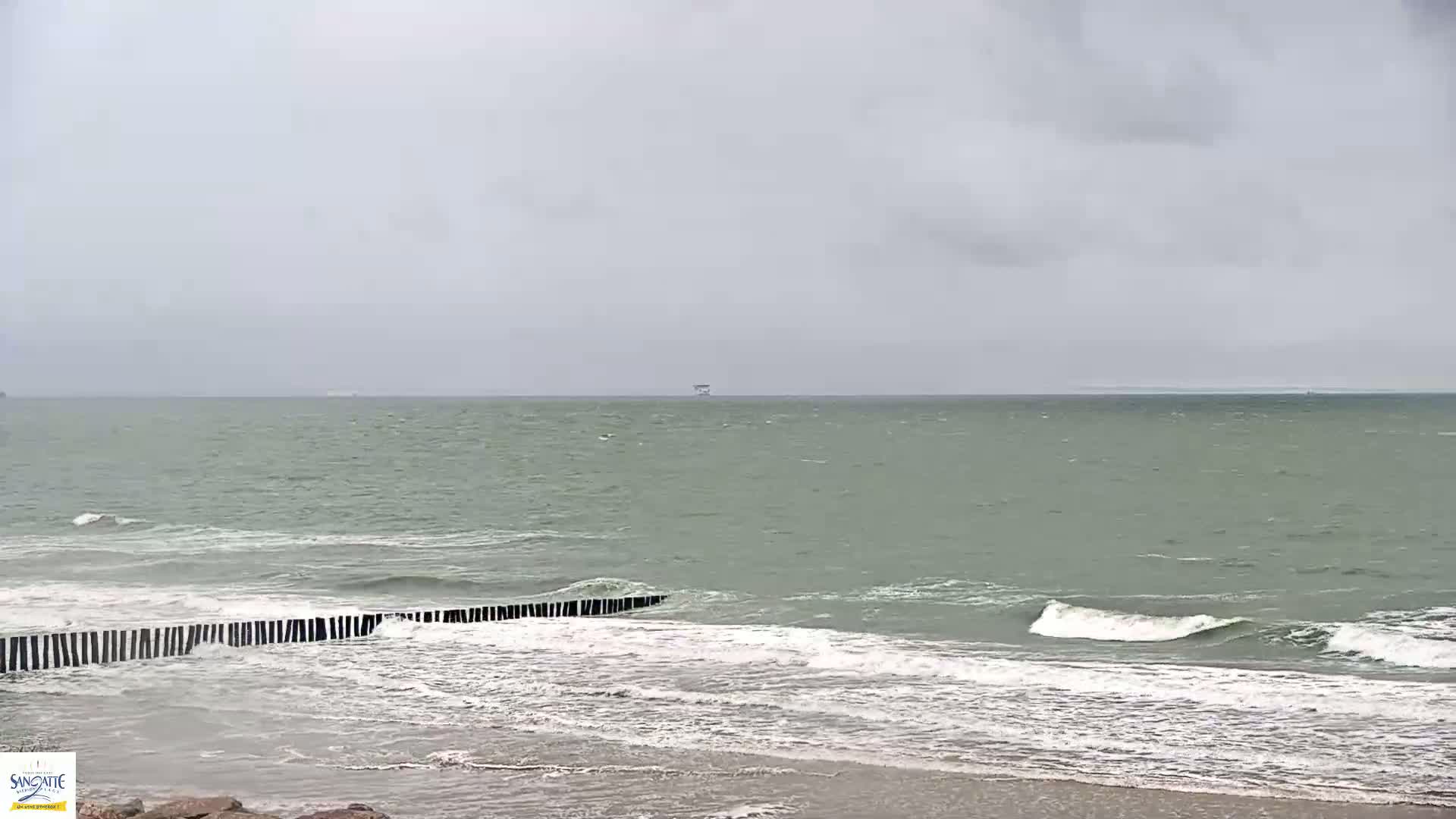 A grey, overcast sky hangs over a choppy sea meeting a sandy beach protected by a row of dark wooden breakwaters.