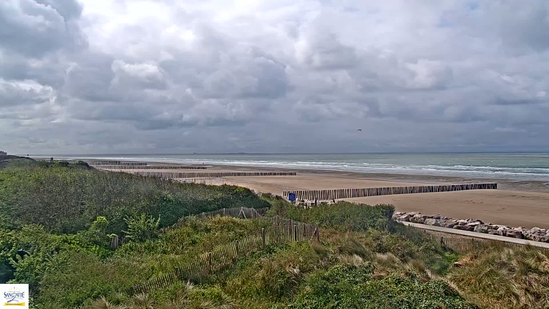 A sandy beach with a wavy ocean under a cloudy sky is partially protected by wooden barriers.