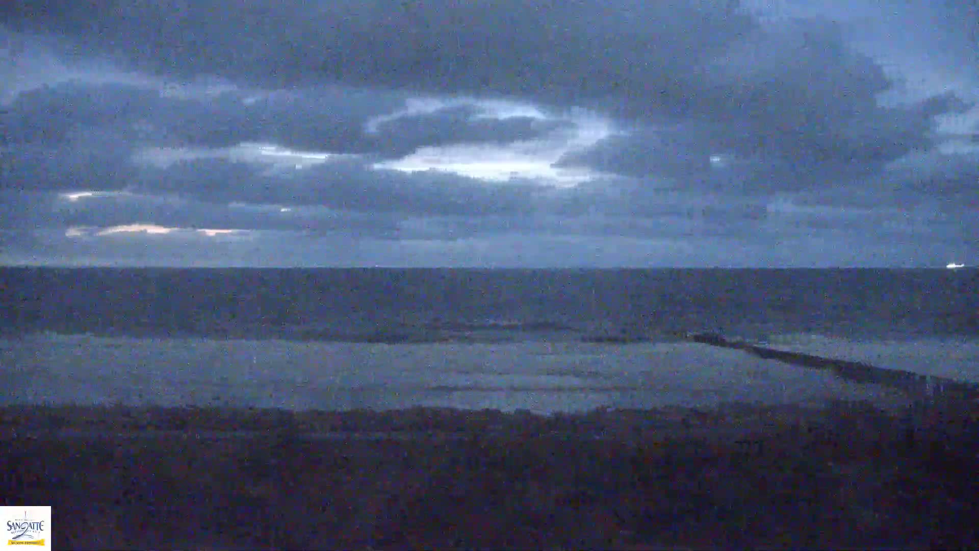 A dark, cloudy sky hangs over a calm, gray ocean, with a long, dark jetty visible in the foreground.
