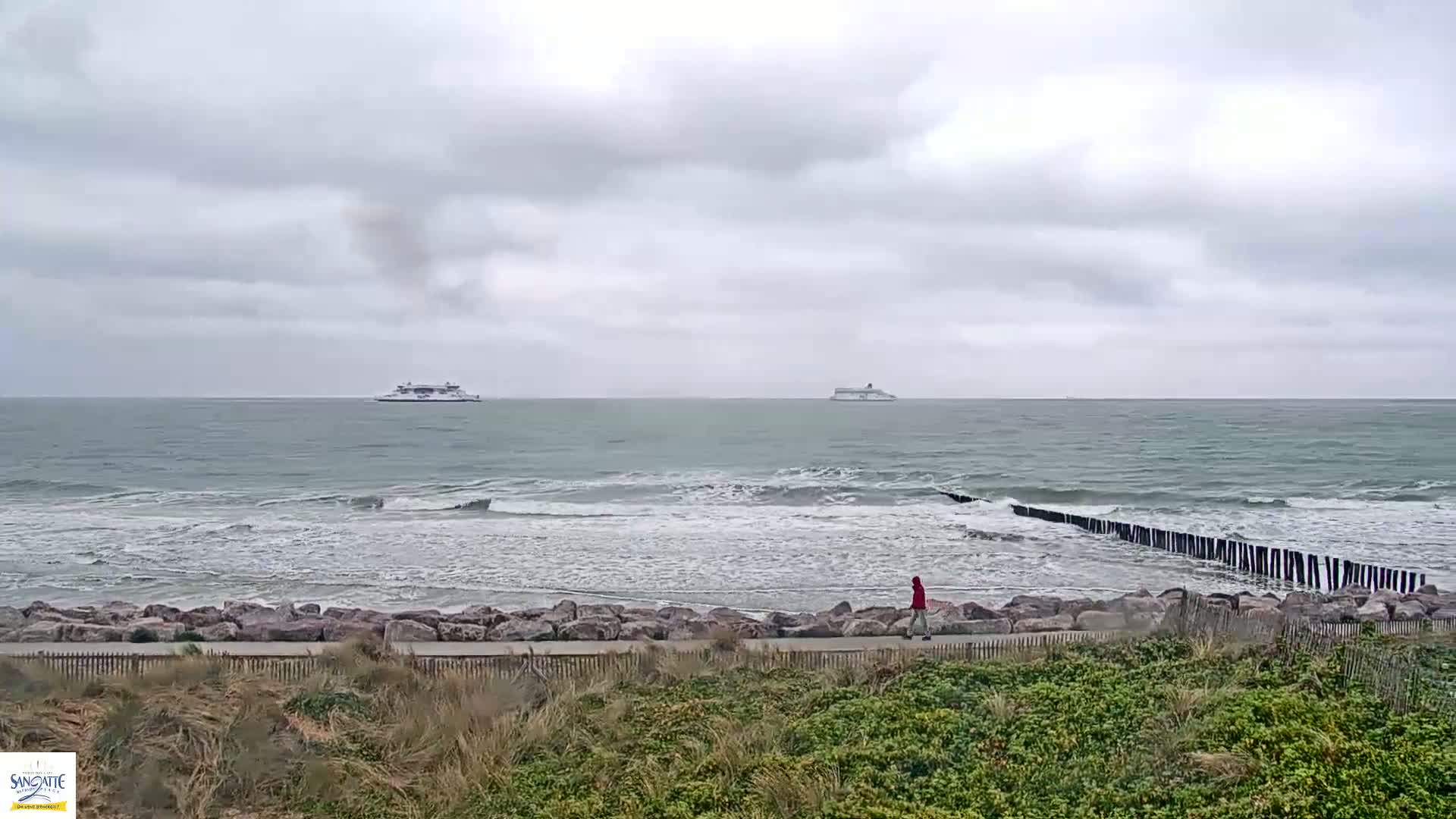A person walks along a rocky shoreline with a breakwater extending into a choppy sea under a cloudy sky, with two ships visible on the horizon.