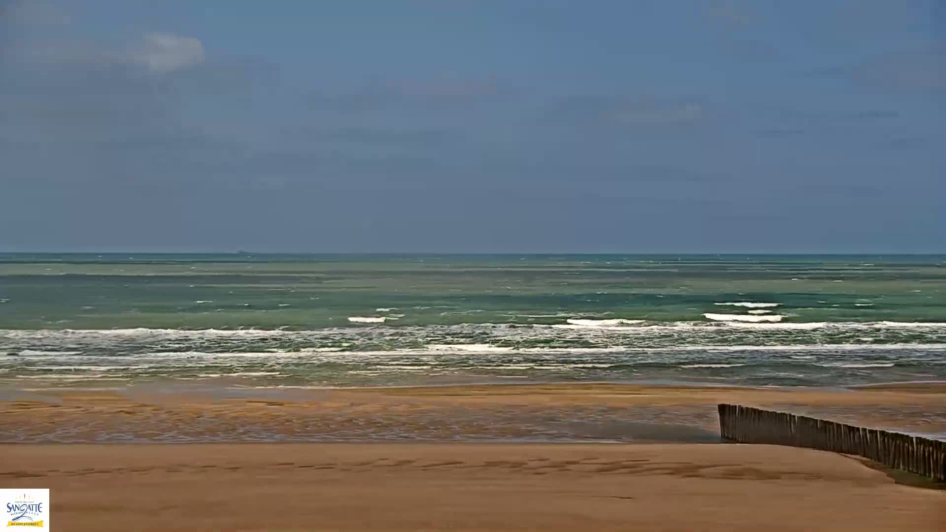 A sandy beach meets a teal ocean with small waves under a mostly clear sky.