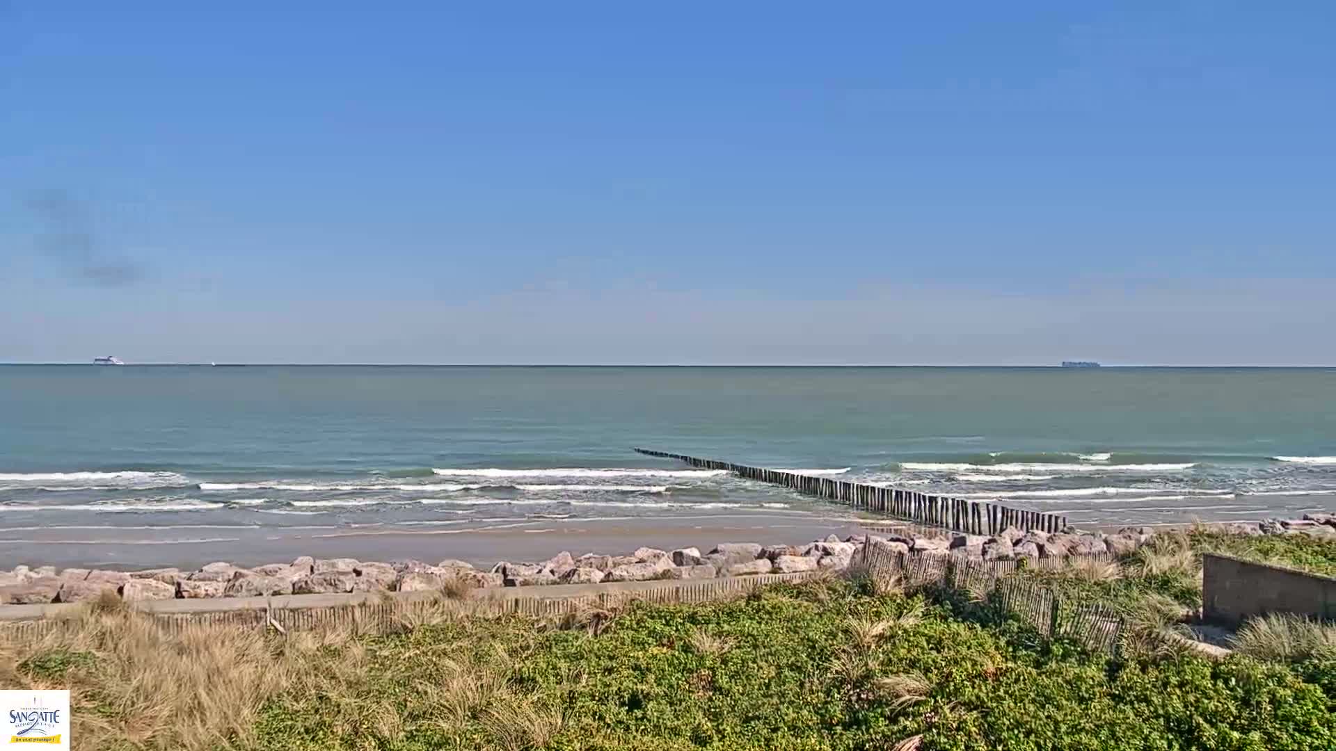 A sandy beach with gentle waves under a clear blue sky, features a rock barrier and wooden posts extending into the ocean.