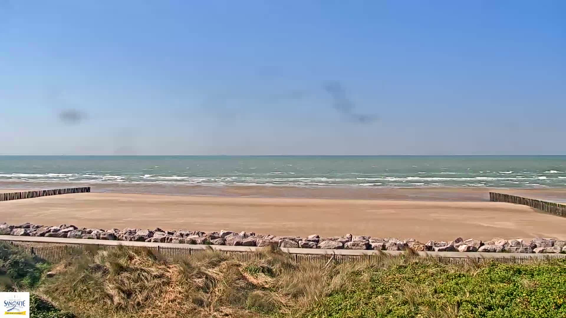 A wide sandy beach with small waves on a calm, sunny day is bordered by rocks and low-lying vegetation.