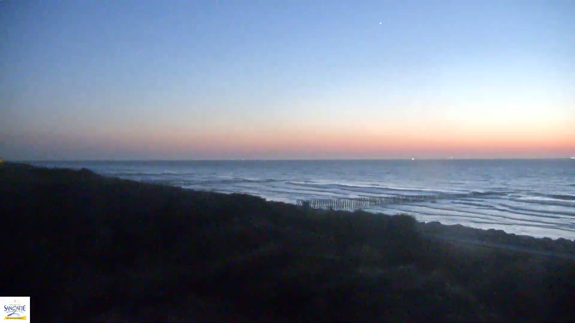 A pink and blue sunset over a dark, wavy ocean with a low, dark shoreline.