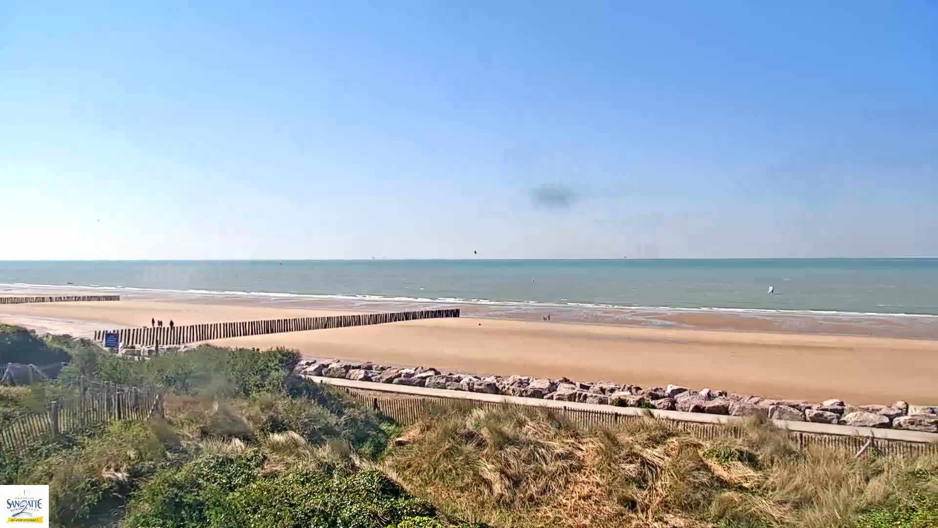 A sandy beach with a calm sea under a clear blue sky is partially protected by wooden posts and a rocky embankment.