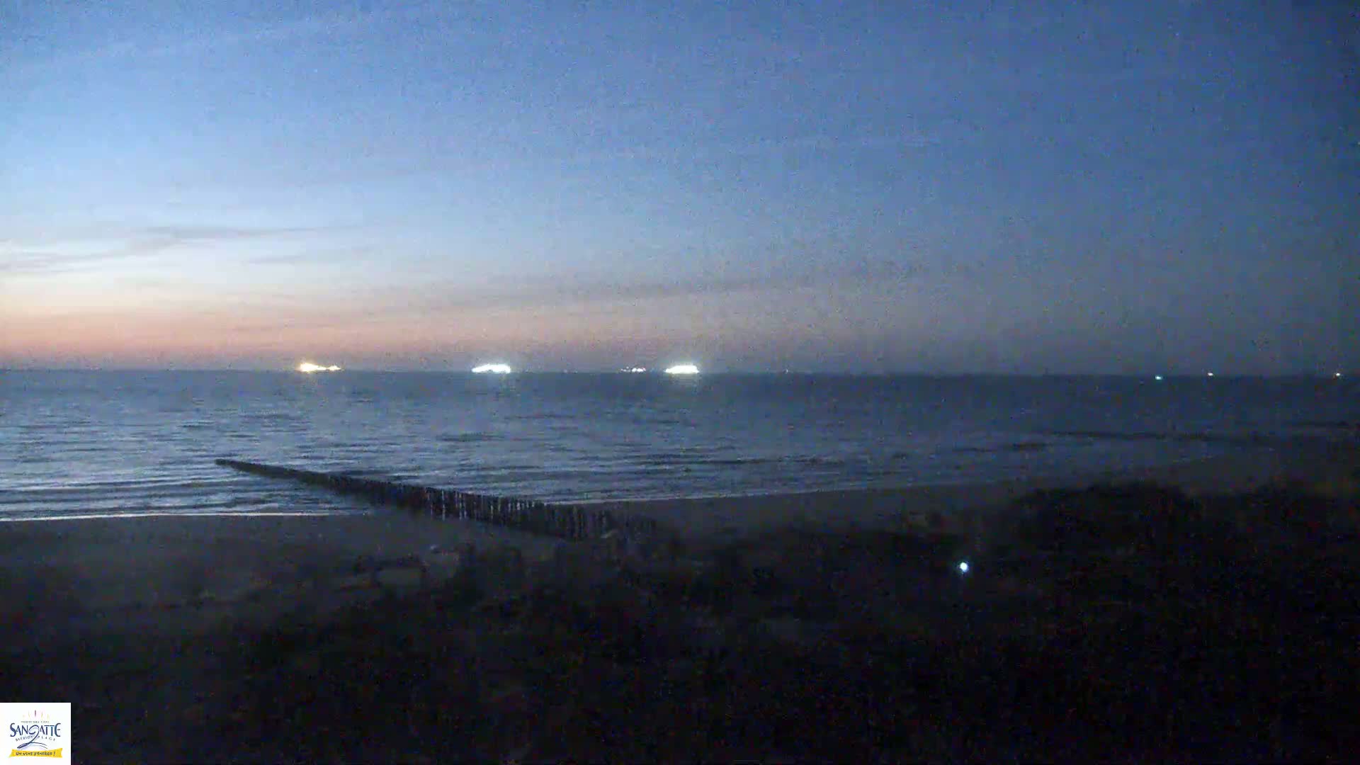 A dark, sandy beach and calm, twilight ocean are visible, with several distant lights on ships at sea.