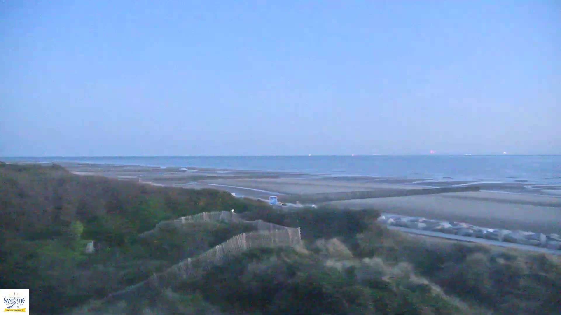 A calm, twilight ocean stretches to the horizon behind a sandy beach and dune-covered landscape.